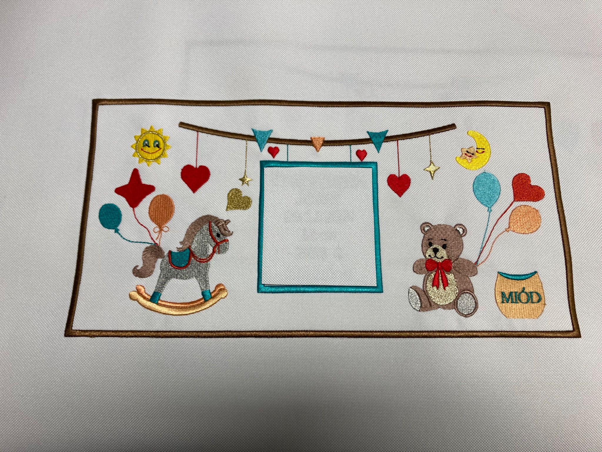 Biała tkanina z haftowanym motywem dziecięcym: konik na biegunach, miś z miodem, słońce, księżyc, balony i girlanda. Puste pole w ramce na zdjęcie.