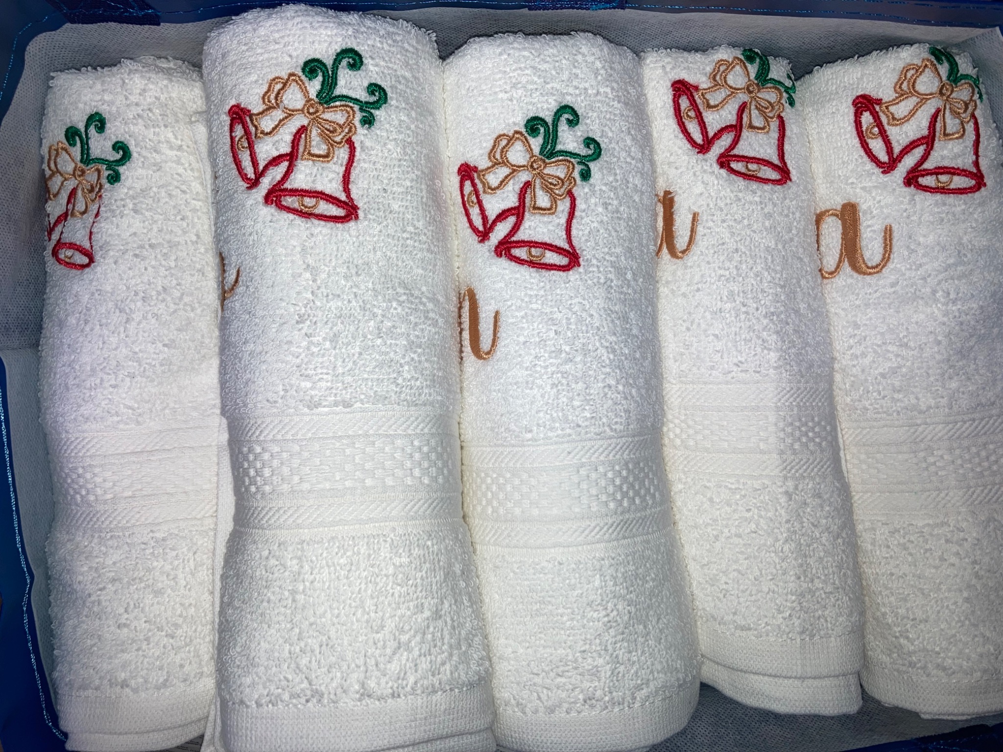 Białe, zwinięte ręczniki z świątecznym motywem dzwonków i inicjałami, zapakowane w pudełko. Dekoracyjny, świąteczny akcent na ręcznikach.