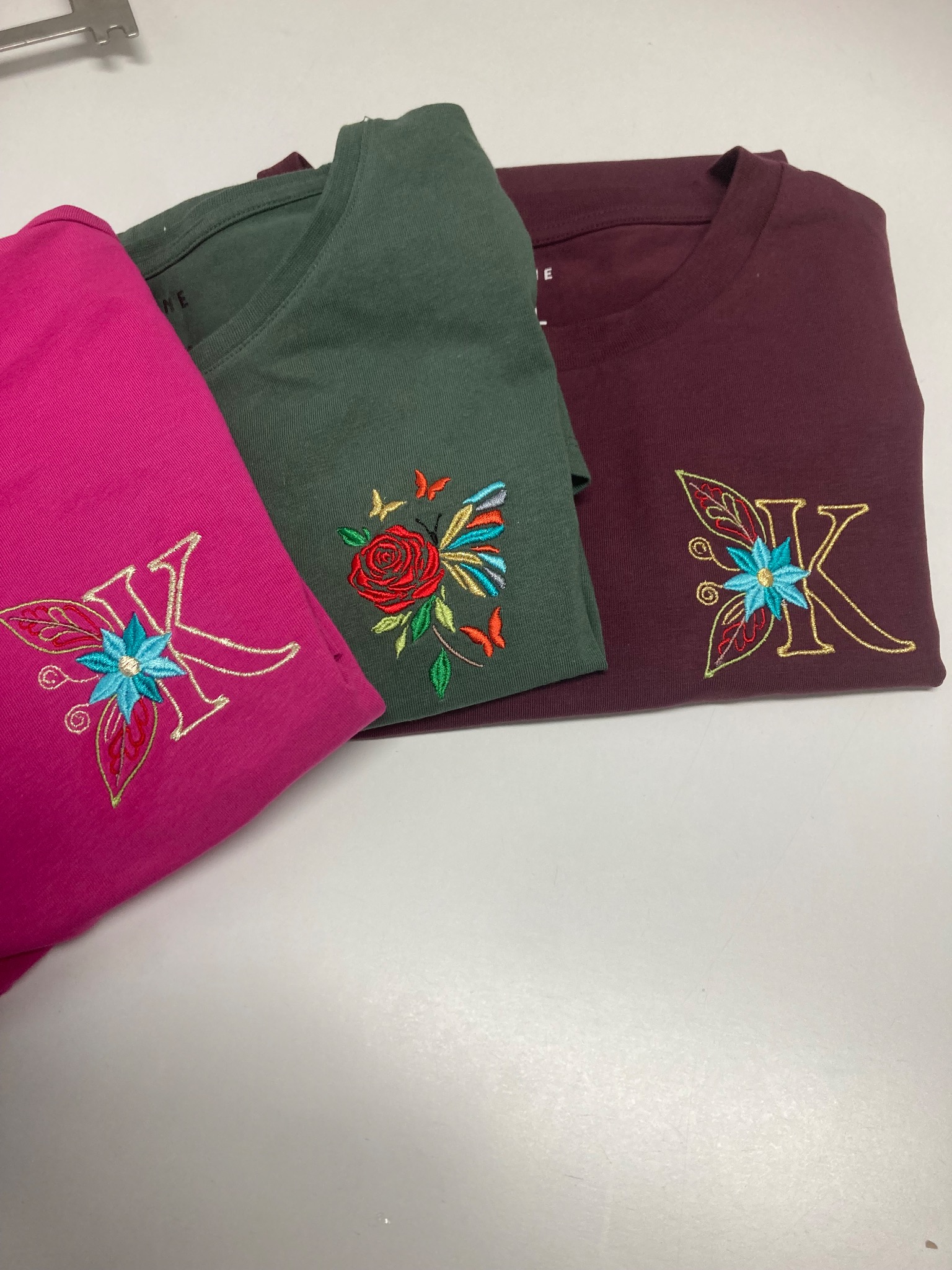 Kolorowe koszulki z motywem kwiatowym i literą K, wykonane w Dębicy. Różowa, zielona i bordowa koszulka z unikalnym wzorem.