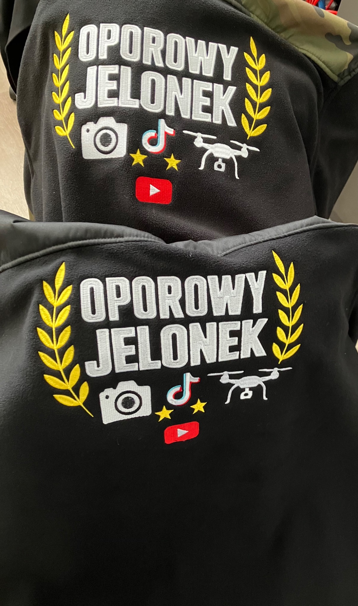Czarna bluza z wyhaftowanym napisem 'OPOROWY JELONEK' otoczonym motywami graficznymi: kamera, TikTok, dron, gwiazdki i YouTube. Detal haftu na odzieży.