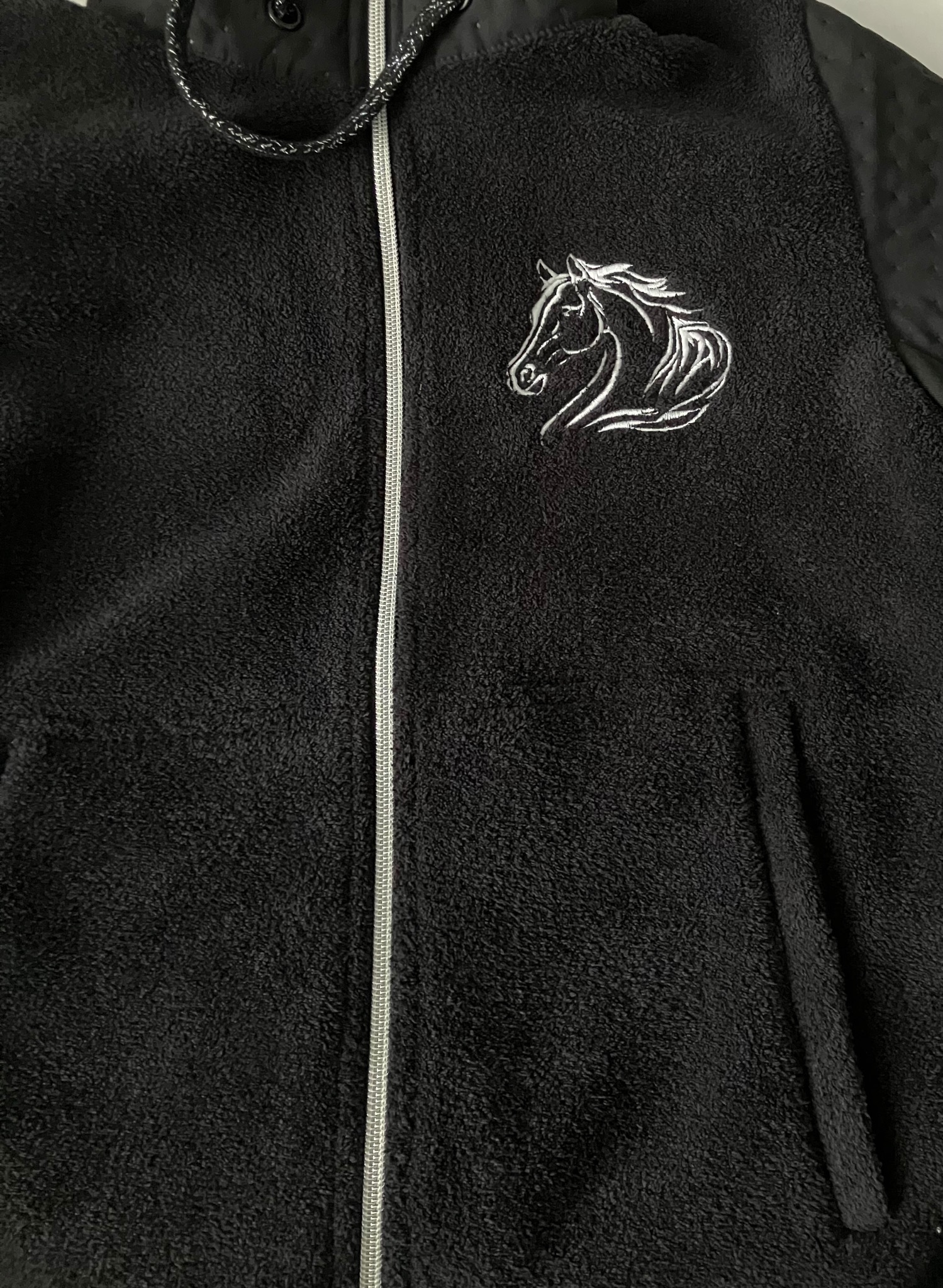 Czarna bluza polarowa z haftowanym białym koniem na piersi, zapinana na zamek błyskawiczny. Widoczny fragment kieszeni i kaptura.