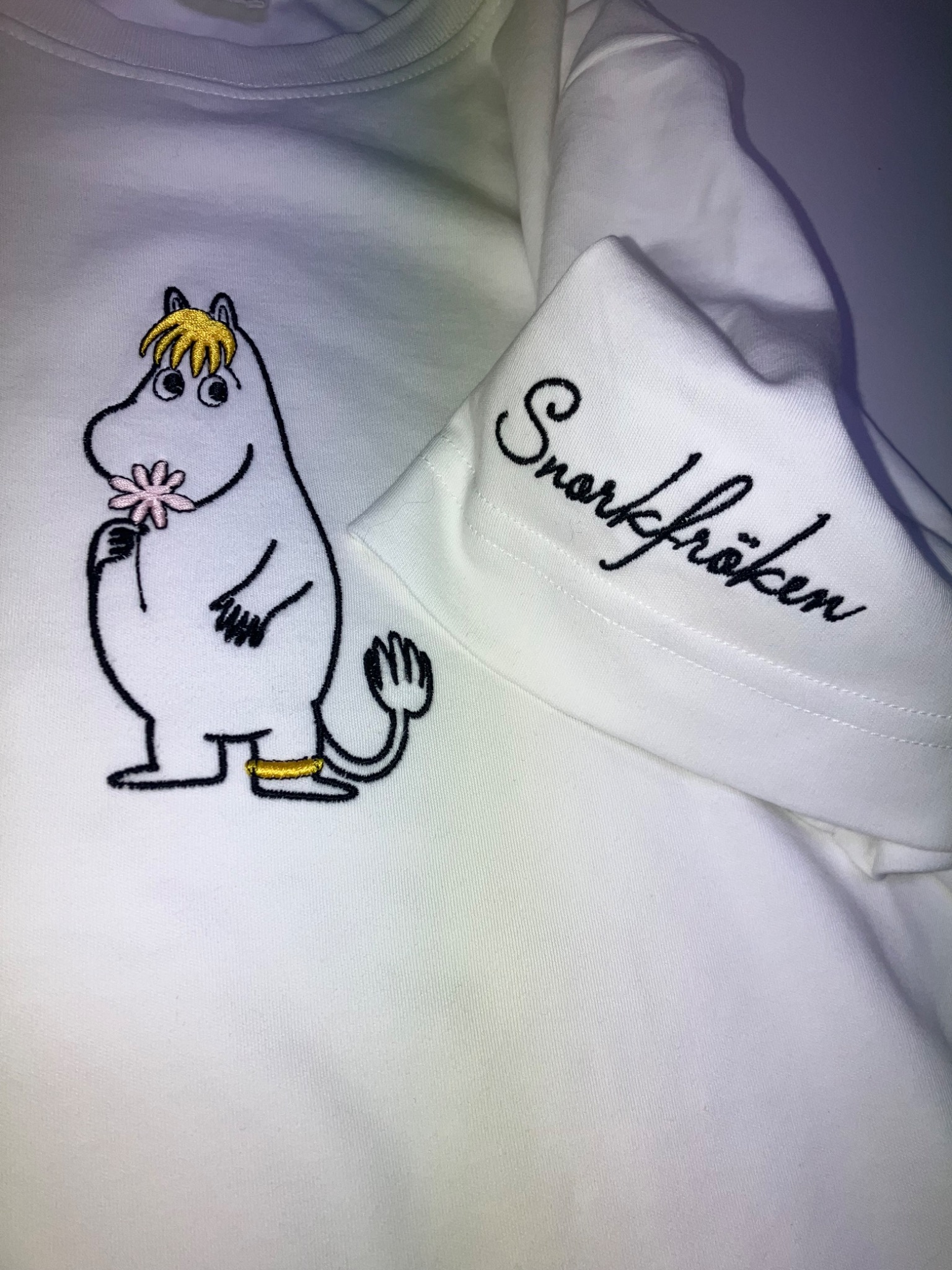 Biała koszulka z haftowanym Muminkiem trzymającym kwiatek i napisem 'Snorkfröken' na rękawie. Detal haftu, bliskie ujęcie.