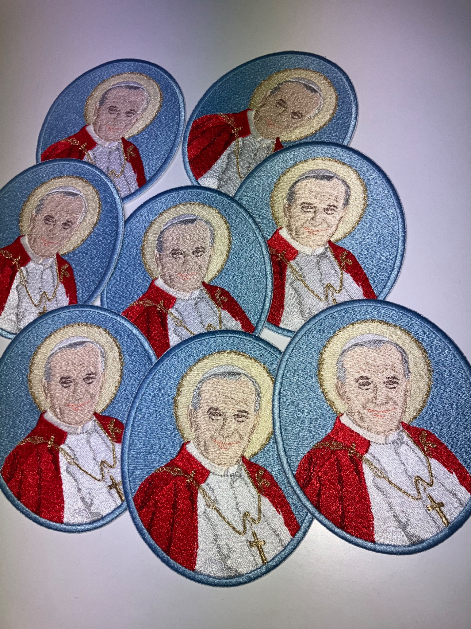 Owalne naszywki z haftowanym portretem papieża Jana Pawła II w czerwonej szacie, otoczonego aureolą, na błękitnym tle. Widoczne detale haftu i złoty łańcuszek z krzyżem.