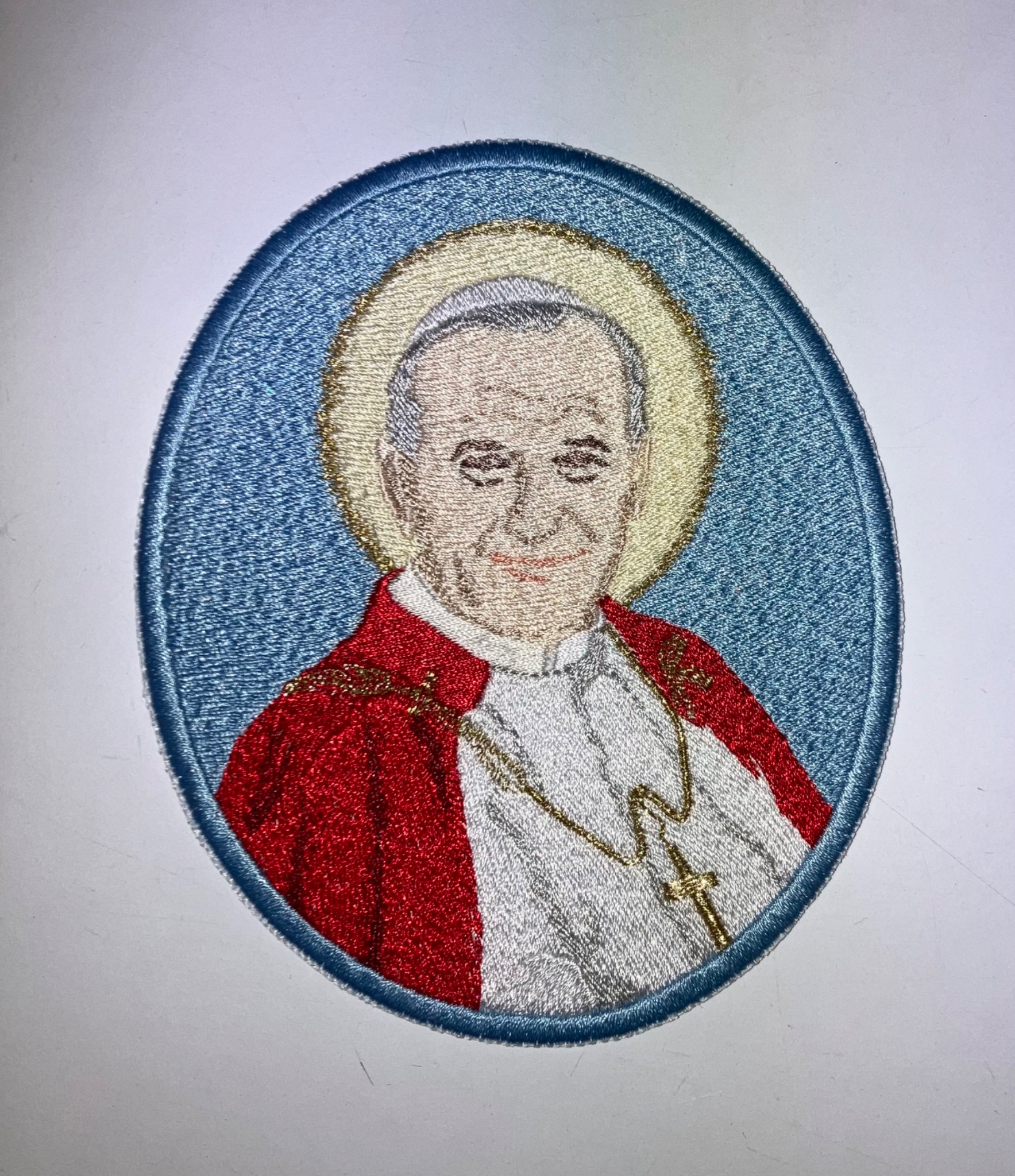 Owalna naszywka z wizerunkiem Jana Pawła II, wykonana techniką haftu maszynowego. Detaliczne odwzorowanie postaci, aureola i szaty w żywych kolorach na jasnym tle.