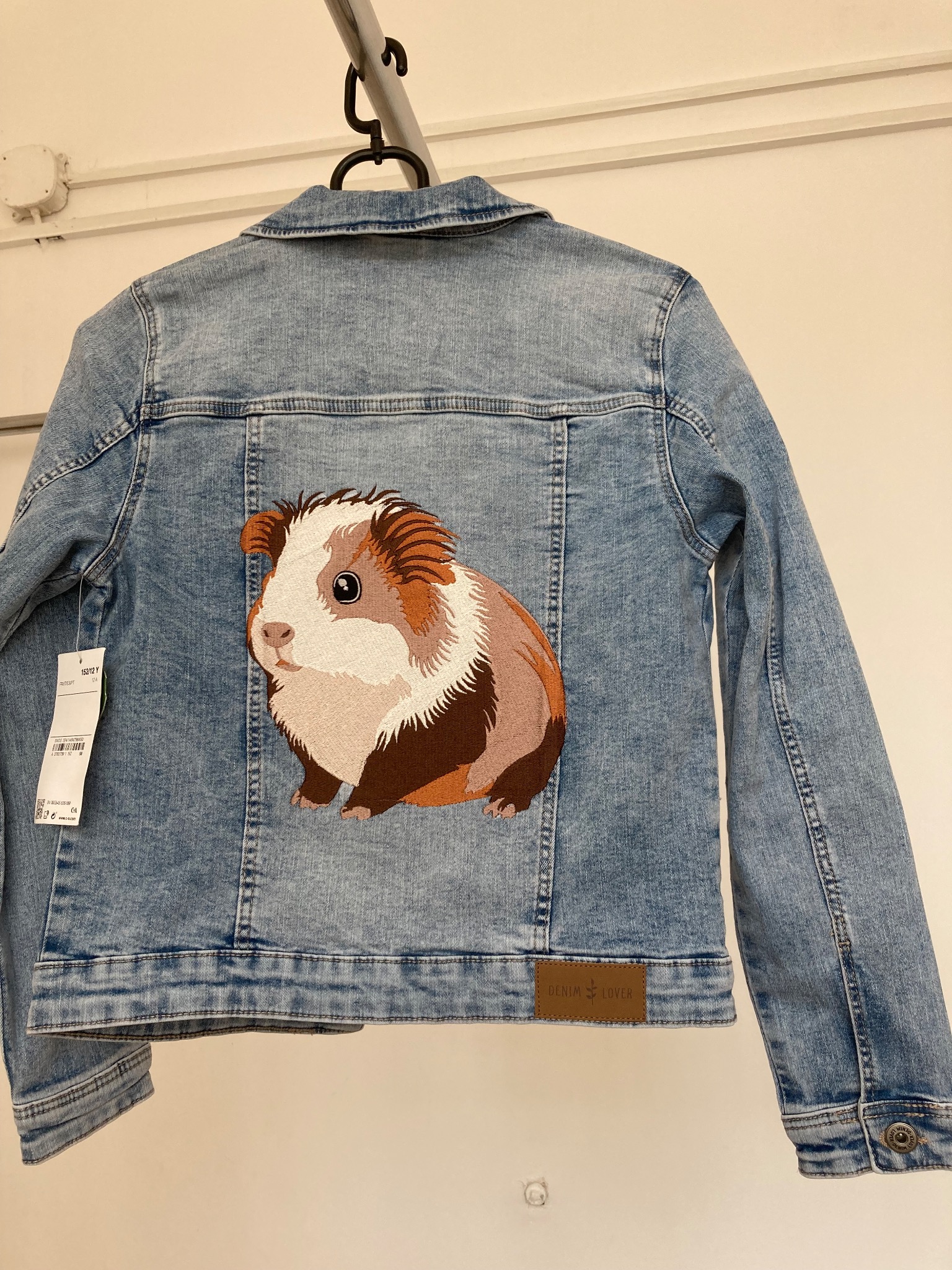 Jasna kurtka jeansowa z haftowanym motywem świnki morskiej na plecach, wisząca na wieszaku.