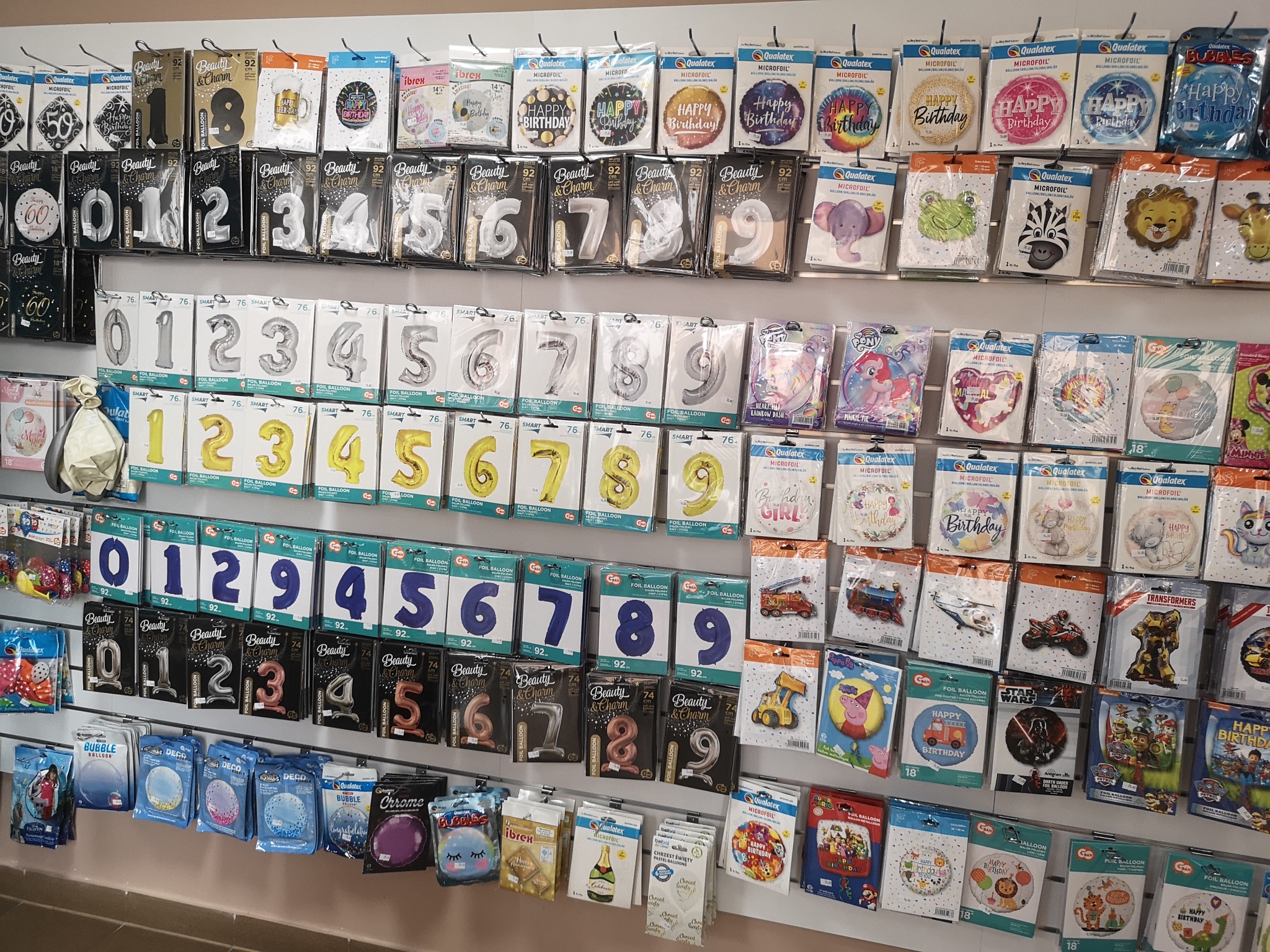 Ekspozycja różnorodnych balonów foliowych i lateksowych w opakowaniach, w tym balony cyfry, balony urodzinowe i balony z postaciami z bajek, rozmieszczonych na półkach sklepowych.