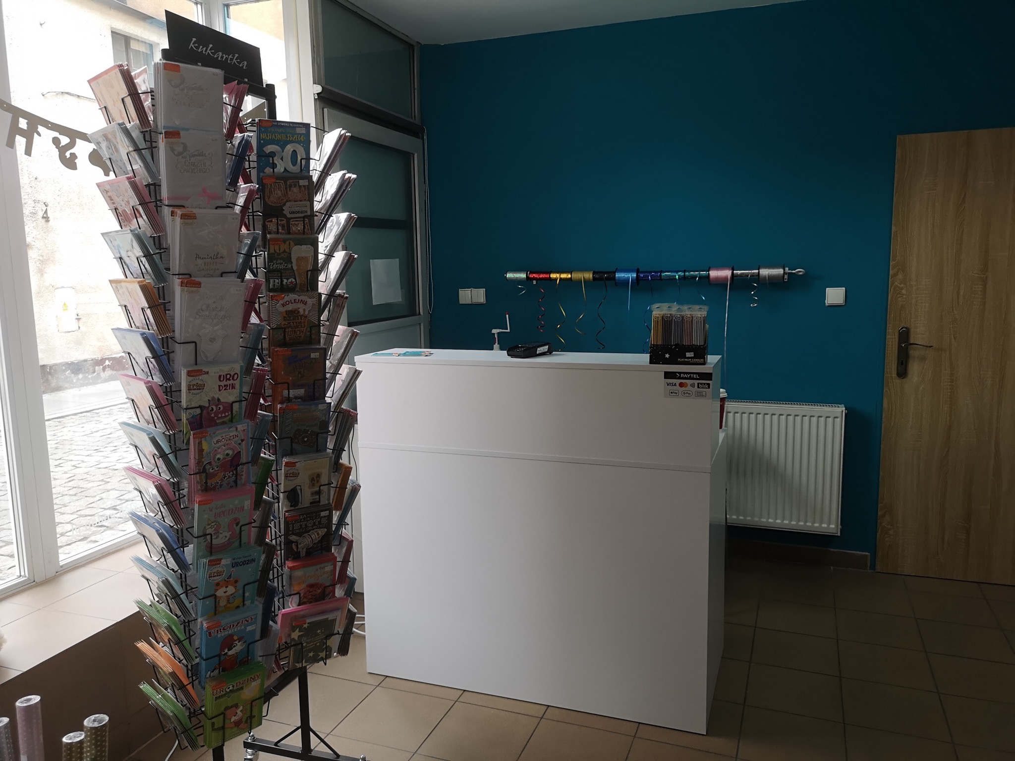 Wnętrze sklepu z kartkami okolicznościowymi: regał z kartkami urodzinowymi i na inne okazje, biała lada recepcyjna, turkusowa ściana z dekoracyjnymi wstążkami.