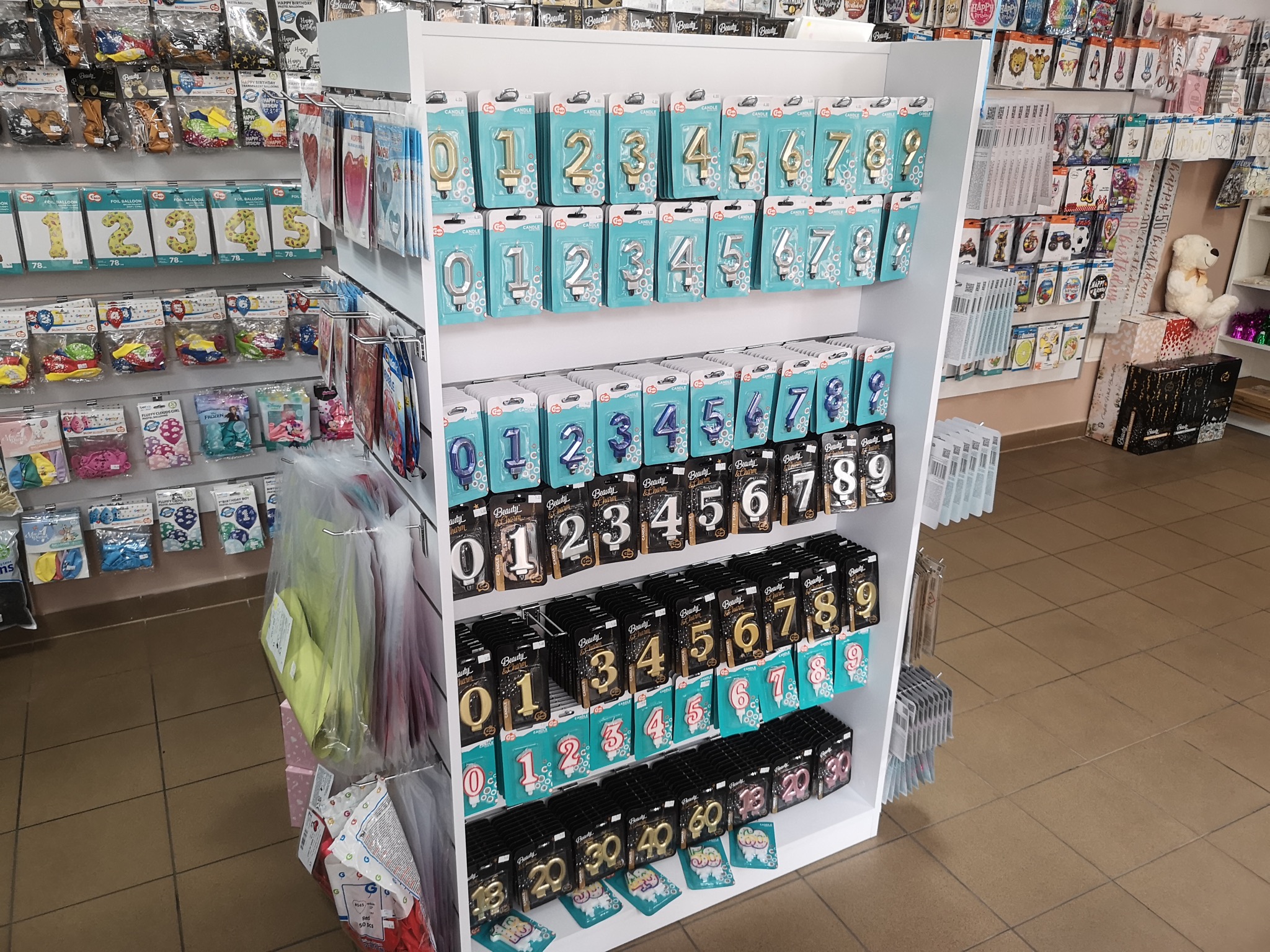 Półka sklepowa z dekoracjami urodzinowymi: świeczki w kształcie cyfr w złotym, srebrnym i niebieskim kolorze, oraz balony foliowe z cyframi, w opakowaniach na tle innych akcesoriów imprezowych.