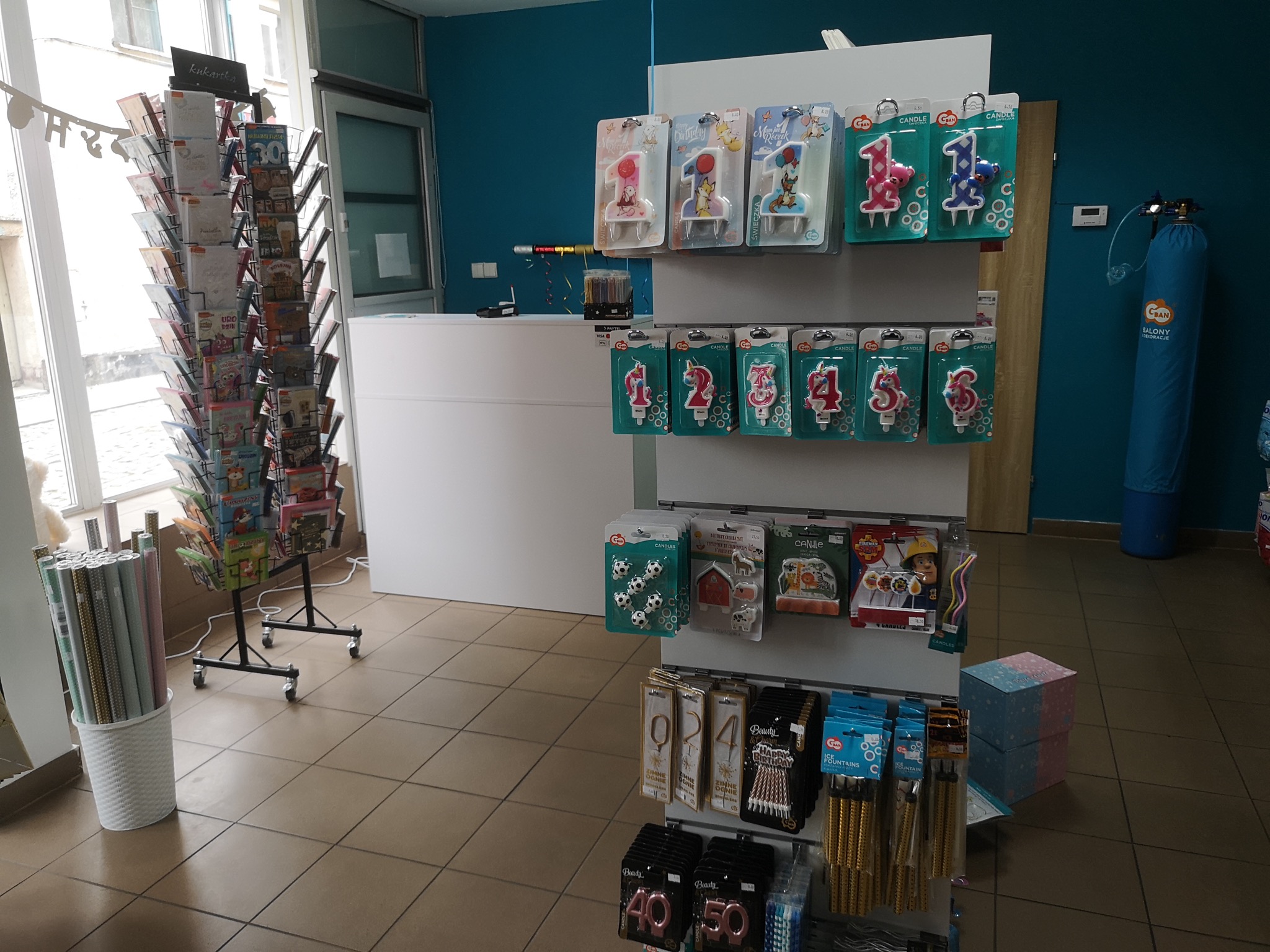 Wnętrze sklepu z artykułami imprezowymi: regały z urodzinowymi świeczkami numerowanymi, fontannami tortowymi, kartkami okolicznościowymi na stojaku i butla z helem w tle.