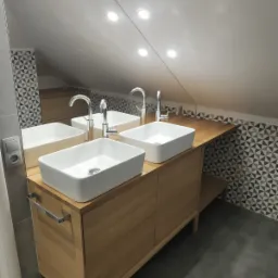 Dwie białe, prostokątne umywalki nablatowe z chromowanymi bateriami, zamontowane na drewnianej szafce w łazience na poddaszu z szarą podłogą i geometryczną mozaiką na ścianie.