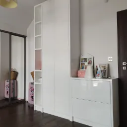 Biała szafa wnękowa z otwartymi półkami i przesuwnymi drzwiami lustrzanymi, uzupełniona komodą z dekoracjami w pokoju dziecięcym z ciemną podłogą.