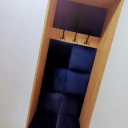 Wnęka w jasnej zabudowie meblowej z drewnianą ramą, wyściełana granatową tapicerką, z trzema metalowymi haczykami na ubrania.