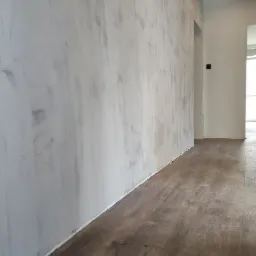 Ściana w trakcie wykańczania z widocznymi smugami szpachli, świeżo położona podłoga z paneli imitujących drewno, widok na korytarz.
