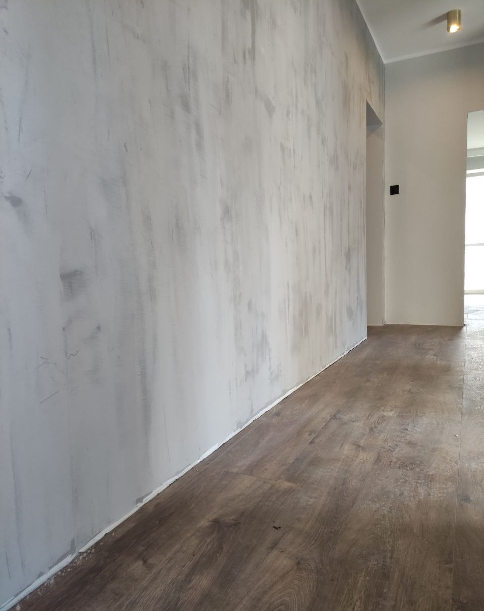 Ściana w trakcie wykańczania z widocznymi smugami szpachli, świeżo położona podłoga z paneli imitujących drewno, widok na korytarz.