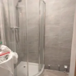 Półokrągła kabina prysznicowa z chromowanymi detalami i szklanymi drzwiami, widoczna na tle szarych płytek imitujących marmur, z elementami zabezpieczającymi podłogę przed zabrudzeniem podczas prac...