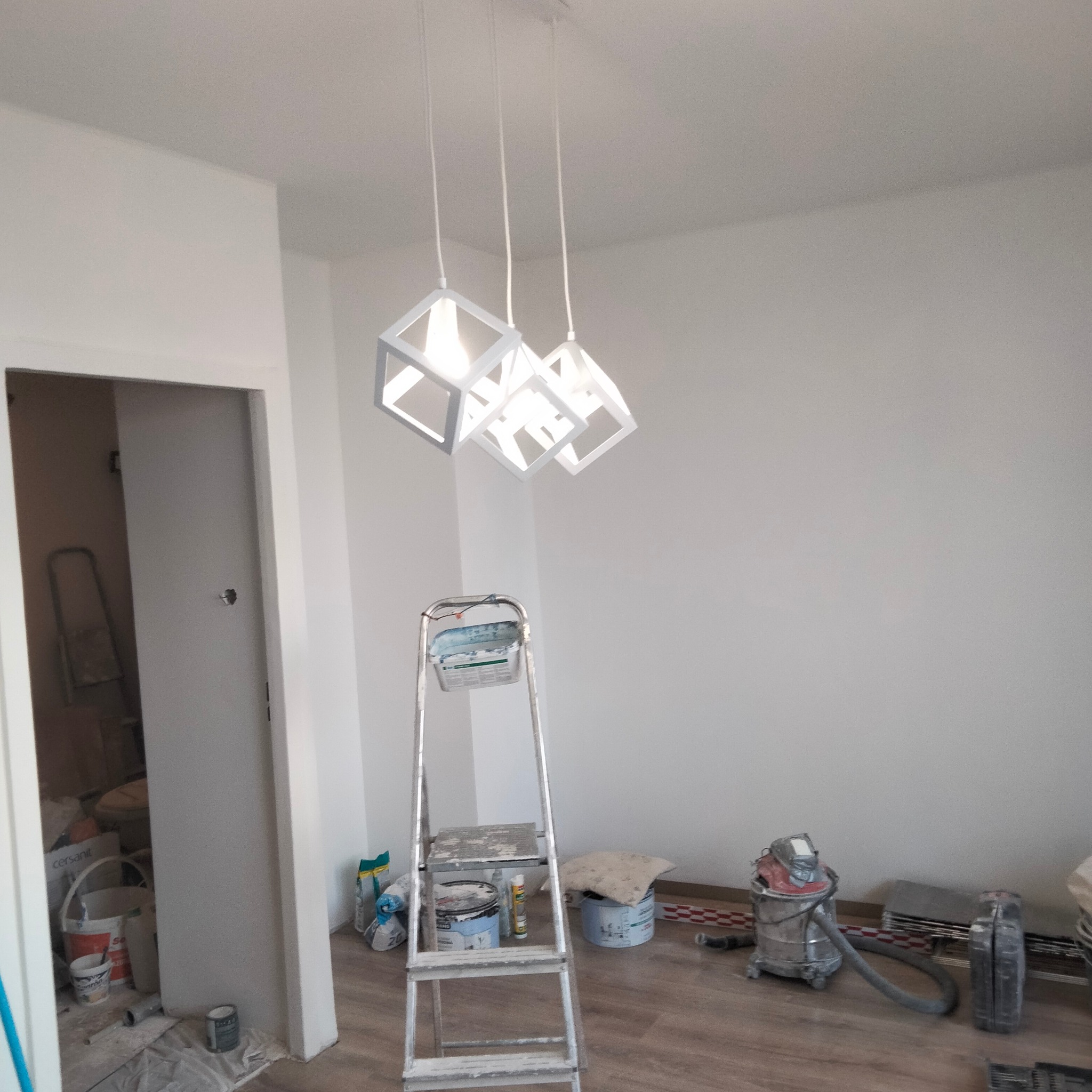 Wnętrze w trakcie malowania ścian na biało, widoczne trzy nowoczesne, białe lampy w kształcie sześcianów, drabina malarska z farbami, odkurzacz przemysłowy i materiały budowlane.