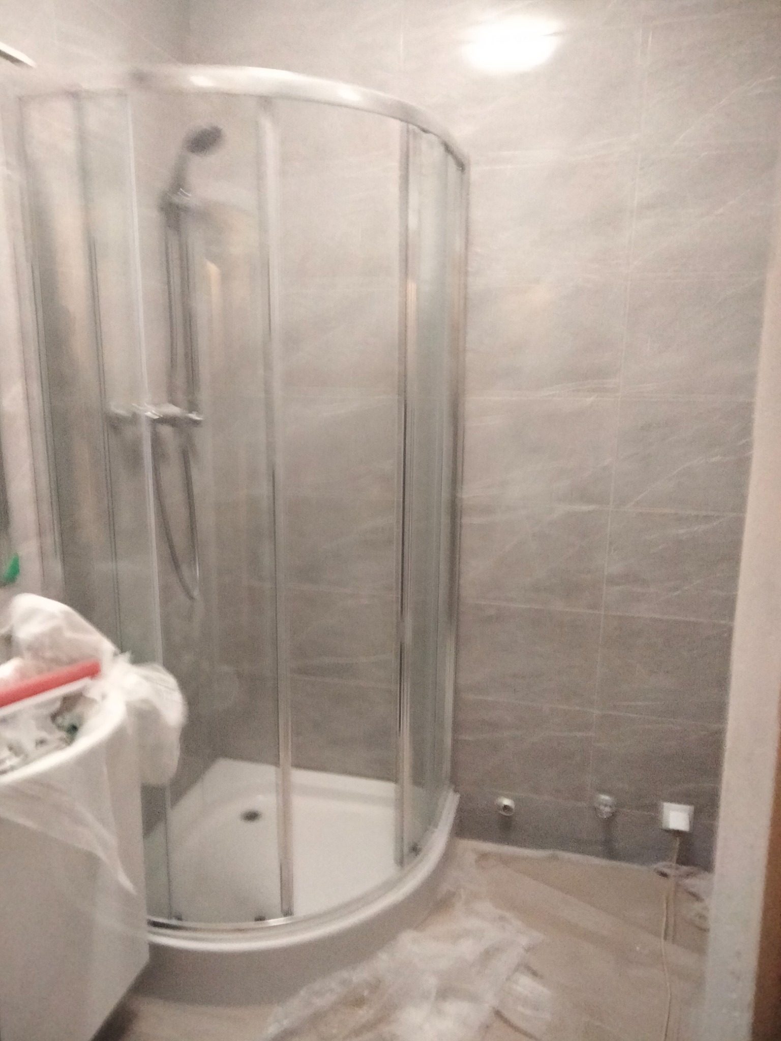 Półokrągła kabina prysznicowa z chromowanymi detalami i szklanymi drzwiami, widoczna na tle szarych płytek imitujących marmur, z elementami zabezpieczającymi podłogę przed zabrudzeniem podczas prac...