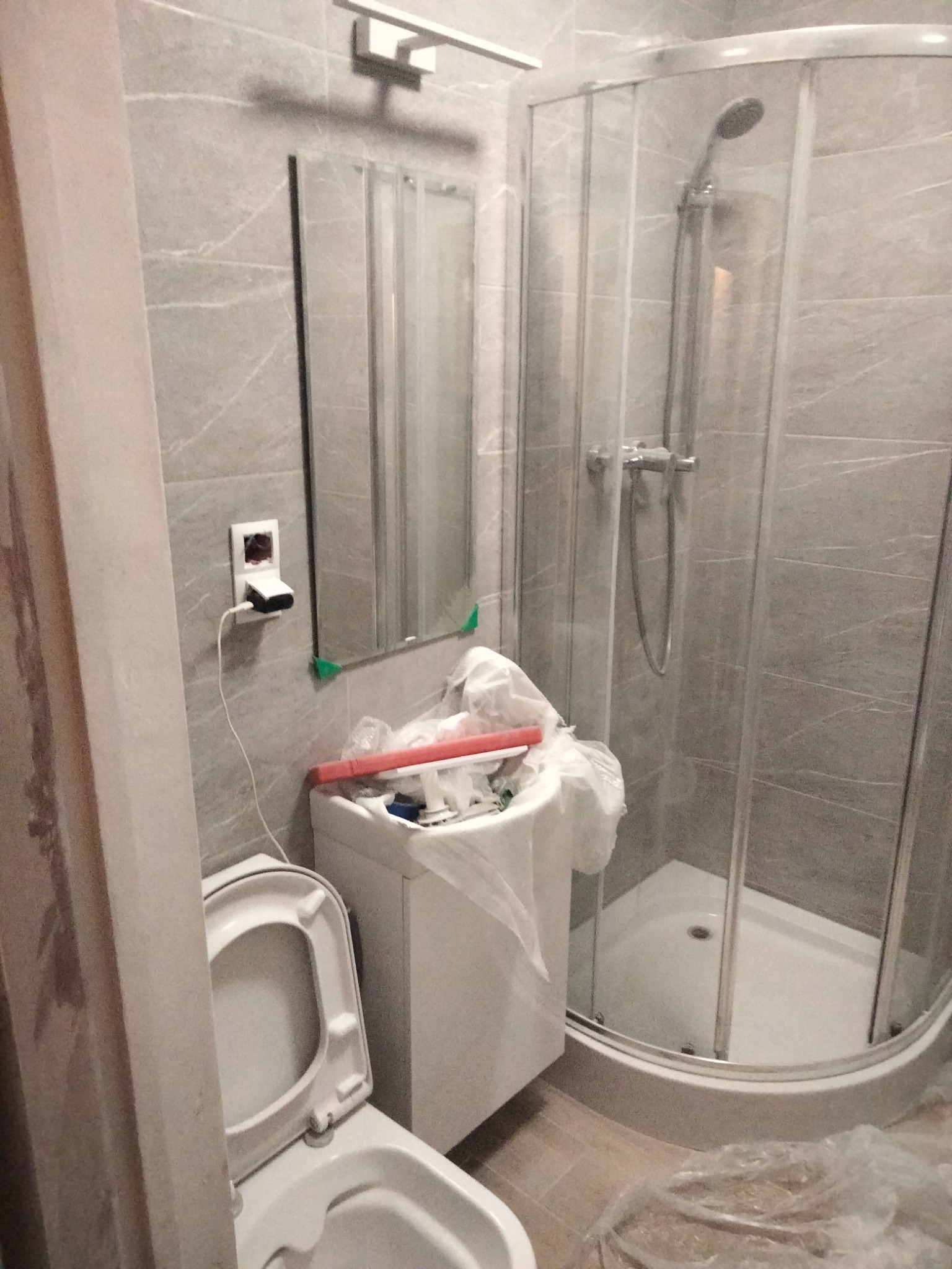 Remont łazienki w trakcie: widoczna kabina prysznicowa z szarymi płytkami, umywalka z narzędziami zabezpieczona folią, częściowo zasłonięta toaleta z otwartą klapą oraz gniazdko elektryczne...