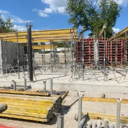 Przygotowanie do wylania fundament&oacute;w z użyciem szalunk&oacute;w systemowych, widoczne zbrojenie i podpory, w tle drzewa i błękitne niebo.