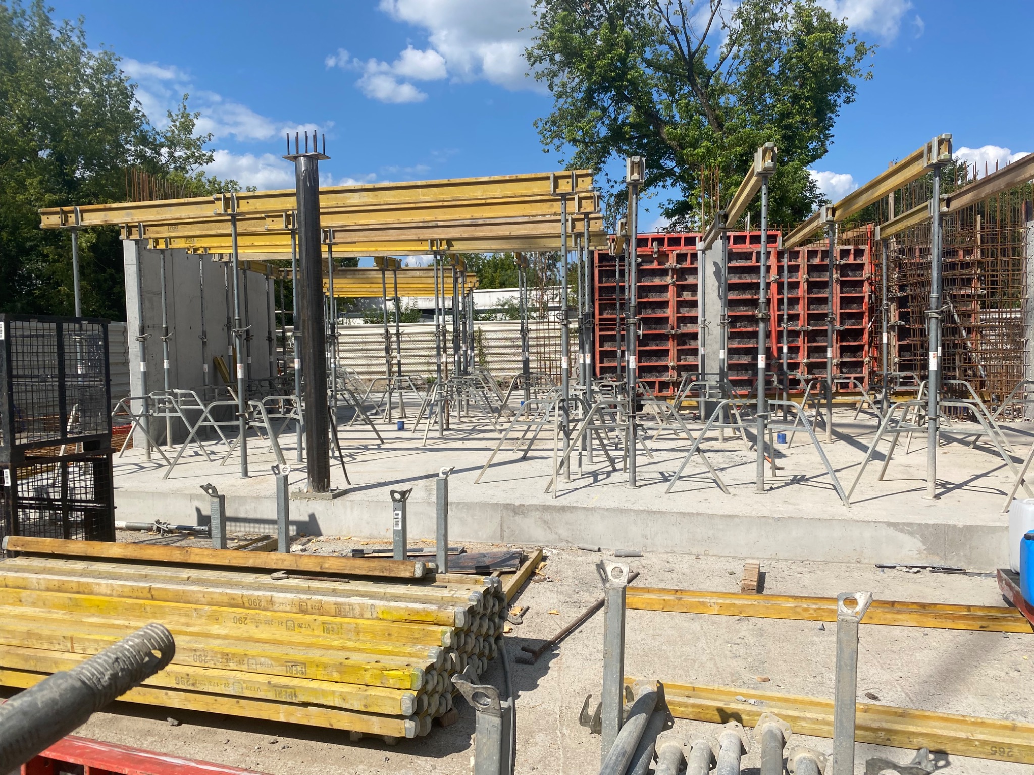 Przygotowanie do wylania fundamentów z użyciem szalunków systemowych, widoczne zbrojenie i podpory, w tle drzewa i błękitne niebo.