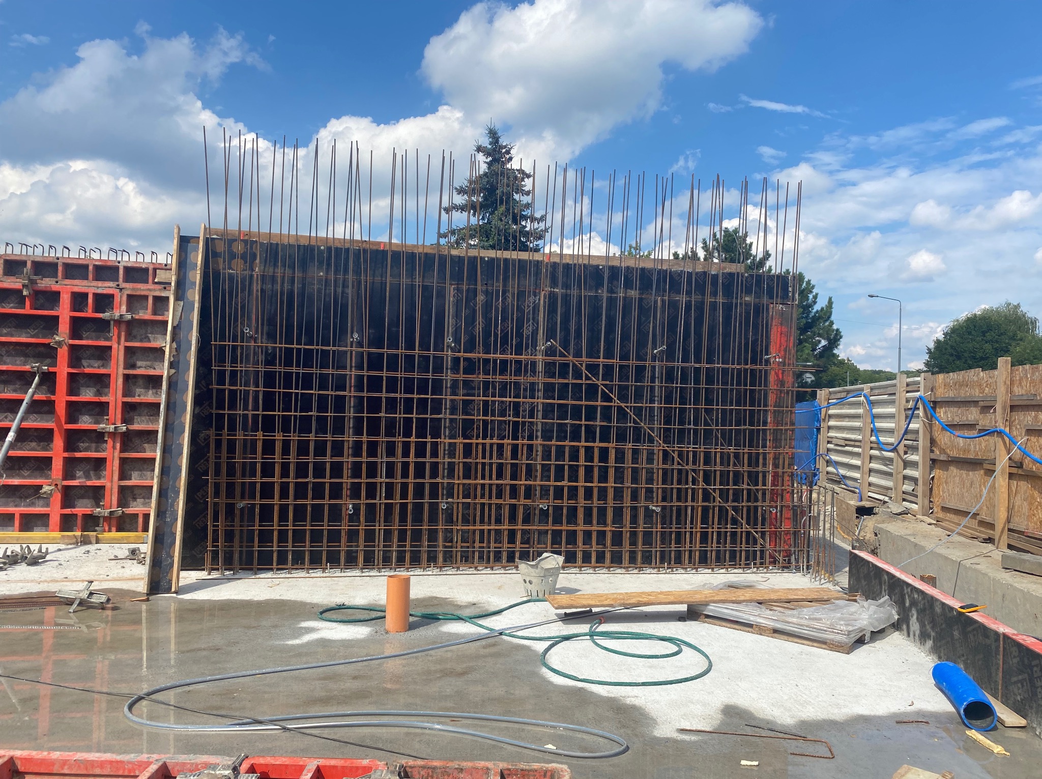 Deskowanie zbrojone stalą, przygotowane do zalania betonem, z widocznymi elementami konstrukcyjnymi i przewodami na mokrym betonowym podłożu, w tle drzewa i błękitne niebo z chmurami.