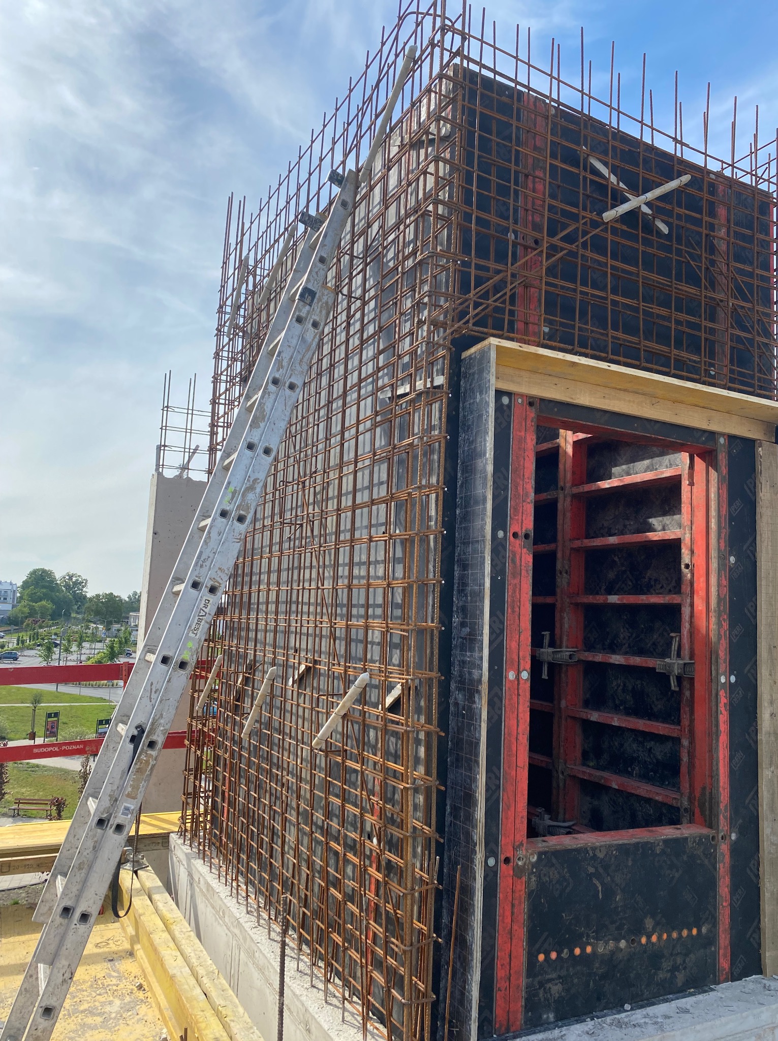 Zbrojenie pionowej ściany budynku w trakcie budowy, z widoczną konstrukcją szalunkową i drabiną opartą o zbrojenie, na tle błękitnego nieba.
