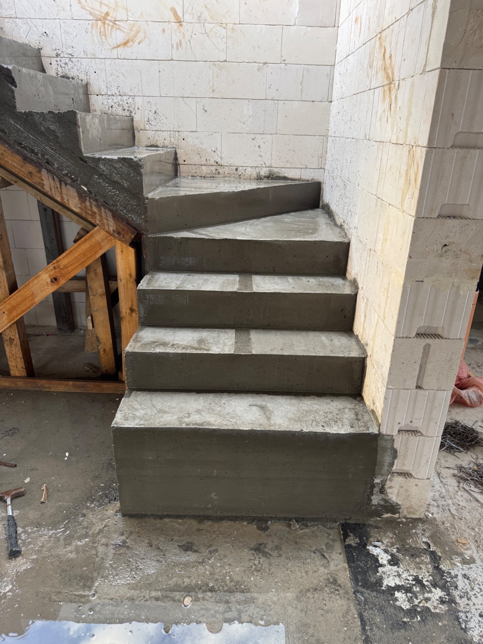 Betonowe schody w surowym stanie, z widocznym drewnianym szalunkiem i murem z bloczków. Ślady wilgoci na betonie i podłożu, hammer leży obok.