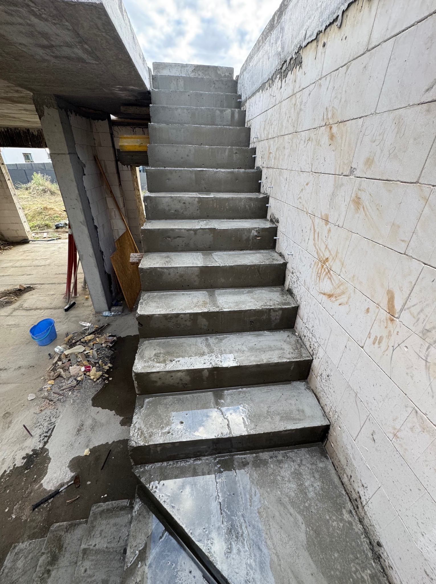 Surowe, betonowe schody w surowym stanie, z widocznymi śladami budowy i elementami konstrukcyjnymi. Ściana z pustaków i niedokończone elementy budynku w tle.