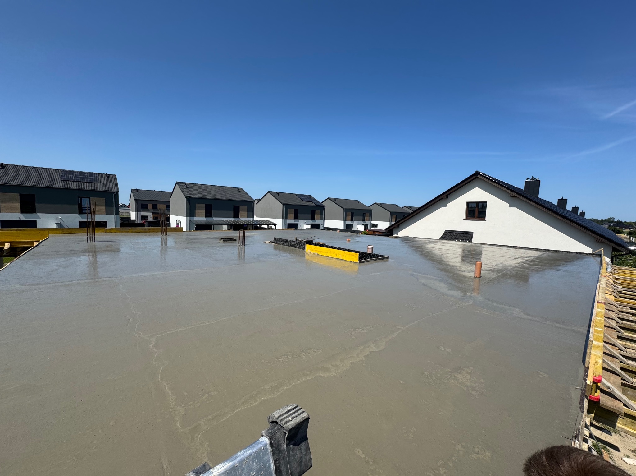 Świeża wylewka na dachu budynku w otoczeniu zabudowy jednorodzinnej, poziomica na powierzchni betonu, błękitne niebo w tle.