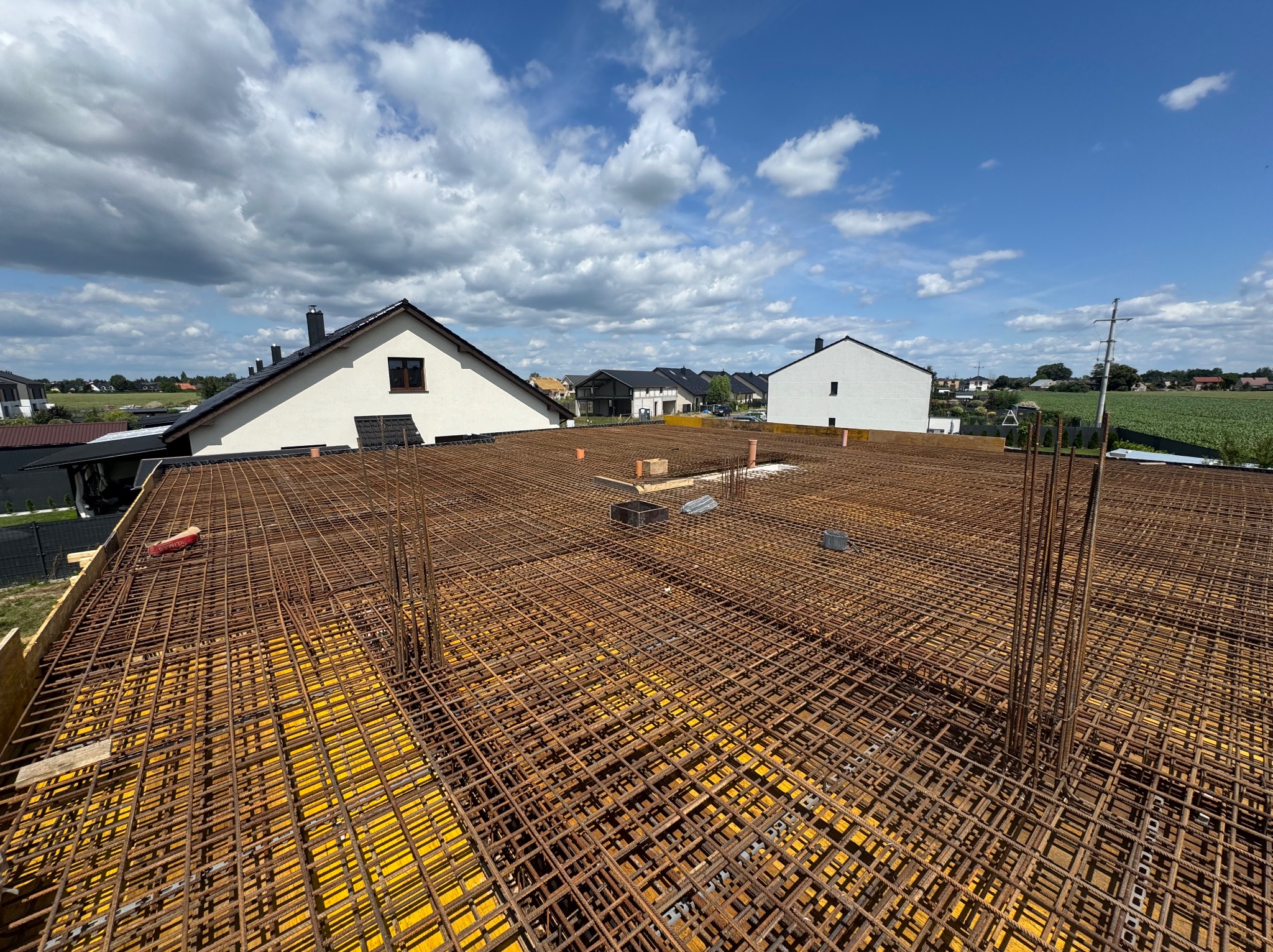 Zbrojenie stropu na żółtym szalunku, widok z góry na konstrukcję z prętów stalowych, w tle budynki mieszkalne i błękitne niebo z chmurami.