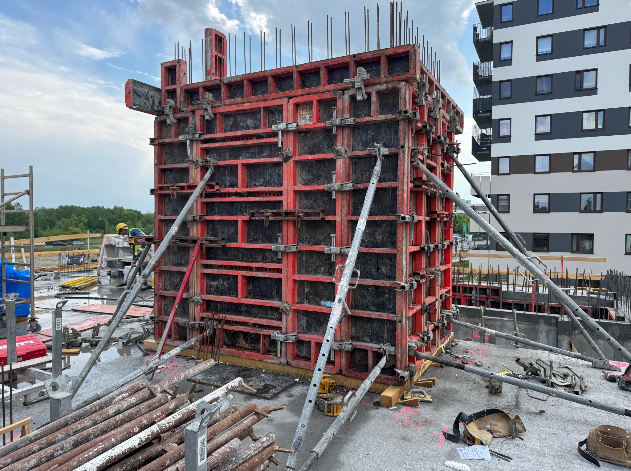 Czerwone szalunki z wystającymi prętami zbrojeniowymi, podparte metalowymi stemplami na budowie z budynkiem w tle, częściowo widoczny pracownik w oddali.