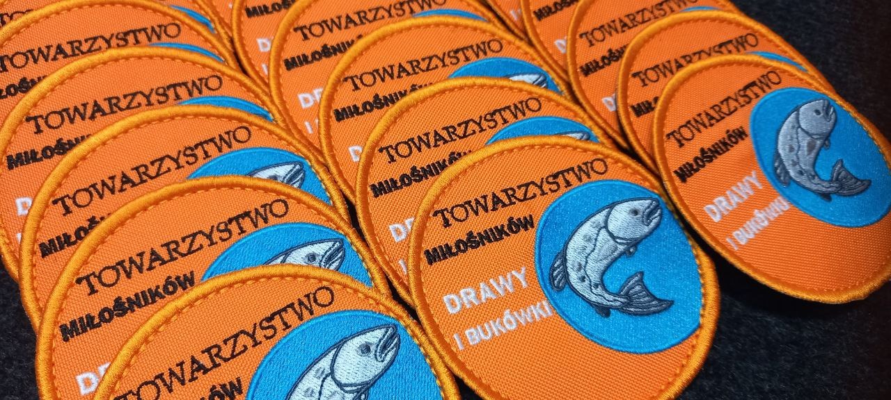 Kolekcja okrągłych naszywek z haftowanym logo przedstawiającym rybę i napisem 'Towarzystwo Miłośników Drawy i Bukówki' na pomarańczowym tle z niebieskim akcentem.
