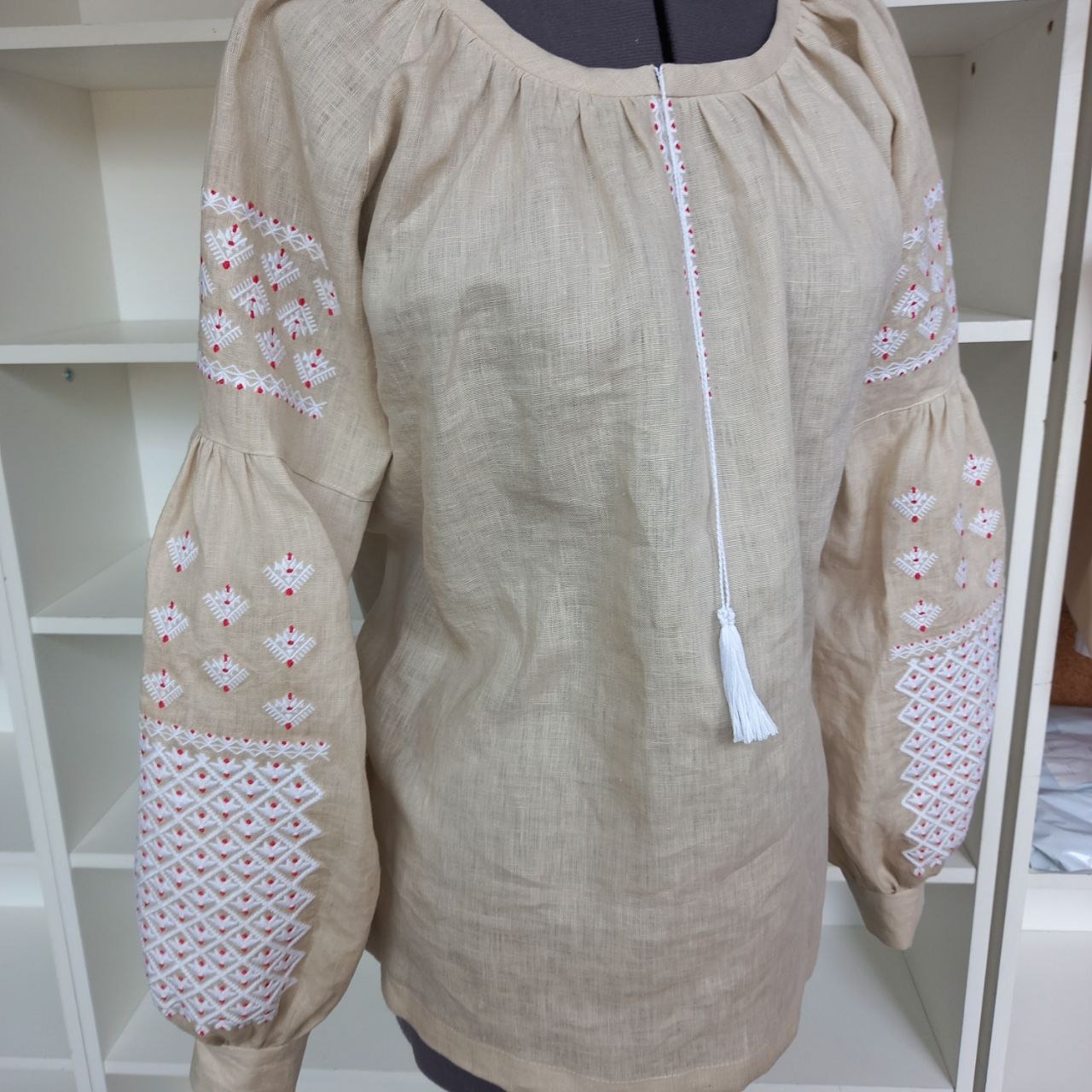 Beżowa lniana bluzka z ręcznie wykonanym białym i czerwonym wzorem geometrycznym na rękawach, widoczna na manekinie na tle białych półek.