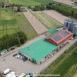 Widok z lotu ptaka na budynek przemysłowy z zielonym dachem i czerwonym dachem z panelami słonecznymi, obok silosy zbożowe, otoczony polami i drzewami.