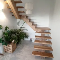 Minimalistyczne, drewniane schody wspornikowe z jasnego drewna, z roślinami doniczkowymi u podstawy, oświetlone wiszącą lampą w kształcie kuli.