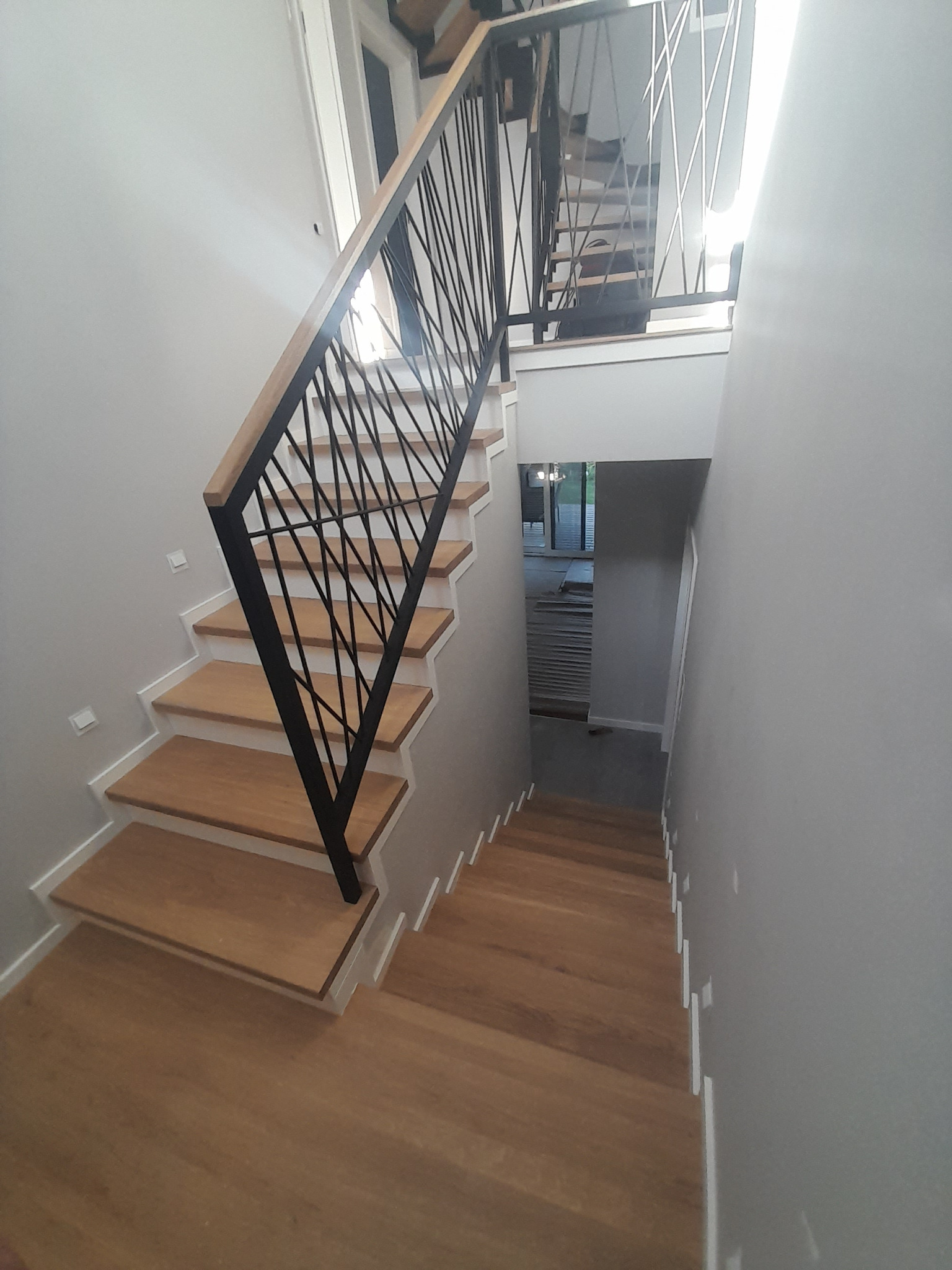 Nowoczesne schody drewniane z czarną metalową balustradą o geometrycznym wzorze, widok z góry w dół, jasne wnętrze z szarymi ścianami.