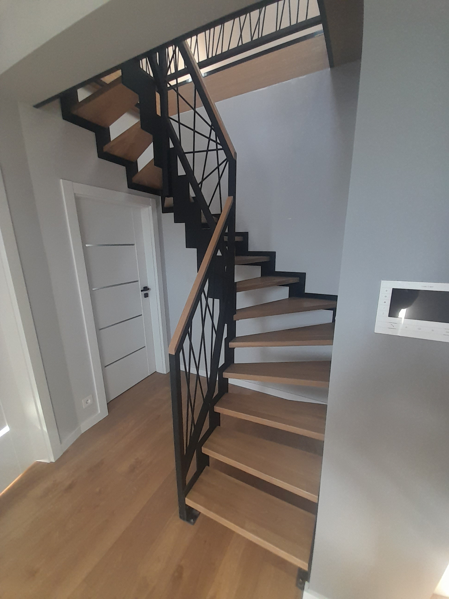 Spiralne schody z drewnianymi stopniami i czarną metalową balustradą z geometrycznym wzorem, widok z dołu, w tle białe drzwi i szara ściana z panelem sterowania.