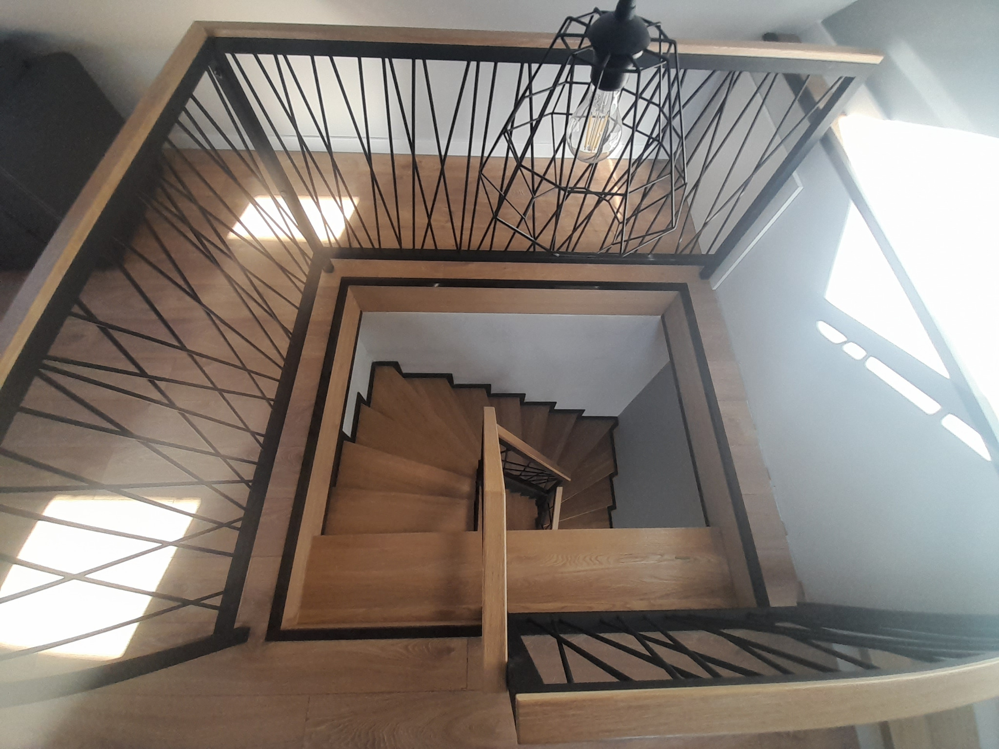 Widok z góry na spiralne, drewniane schody z czarną, metalową balustradą i ozdobną lampą w Lublinie.