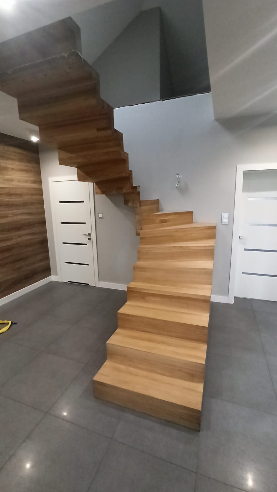 Ażurowe, jednobiegowe schody z jasnego drewna, bez poręczy, zamontowane w nowoczesnym wnętrzu z szarą podłogą i białymi drzwiami.