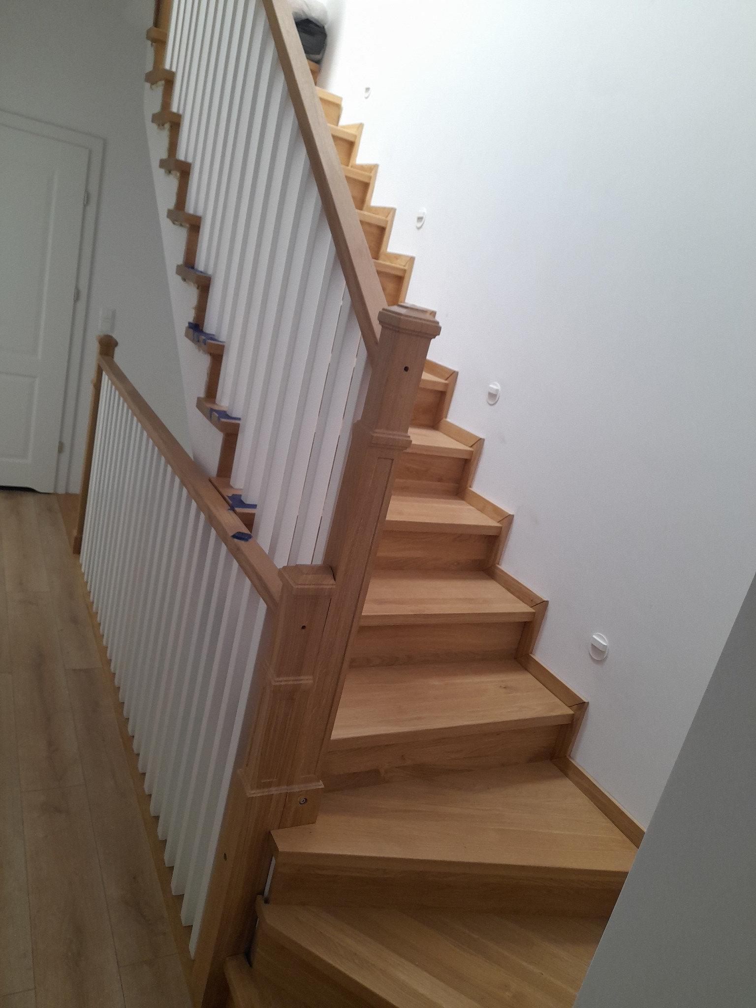 Jasne, drewniane schody z białymi balustradami w nowoczesnym wnętrzu, widoczne ślady niebieskiej taśmy malarskiej zabezpieczającej podczas montażu.