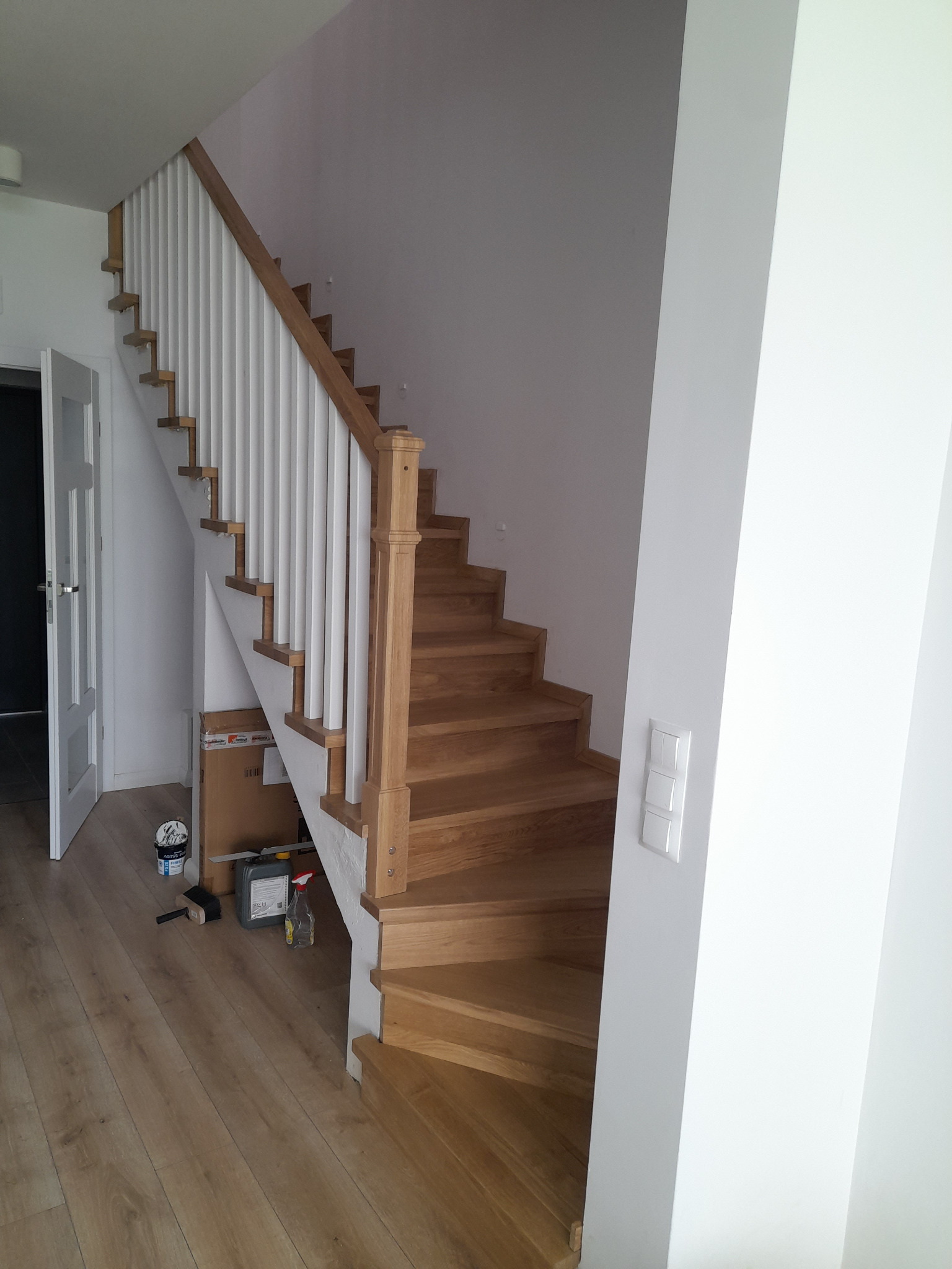Jasne, dębowe stopnie schodów z białymi pionowymi elementami balustrady, widoczne narzędzia i materiały wykończeniowe w pobliżu podstawy schodów.