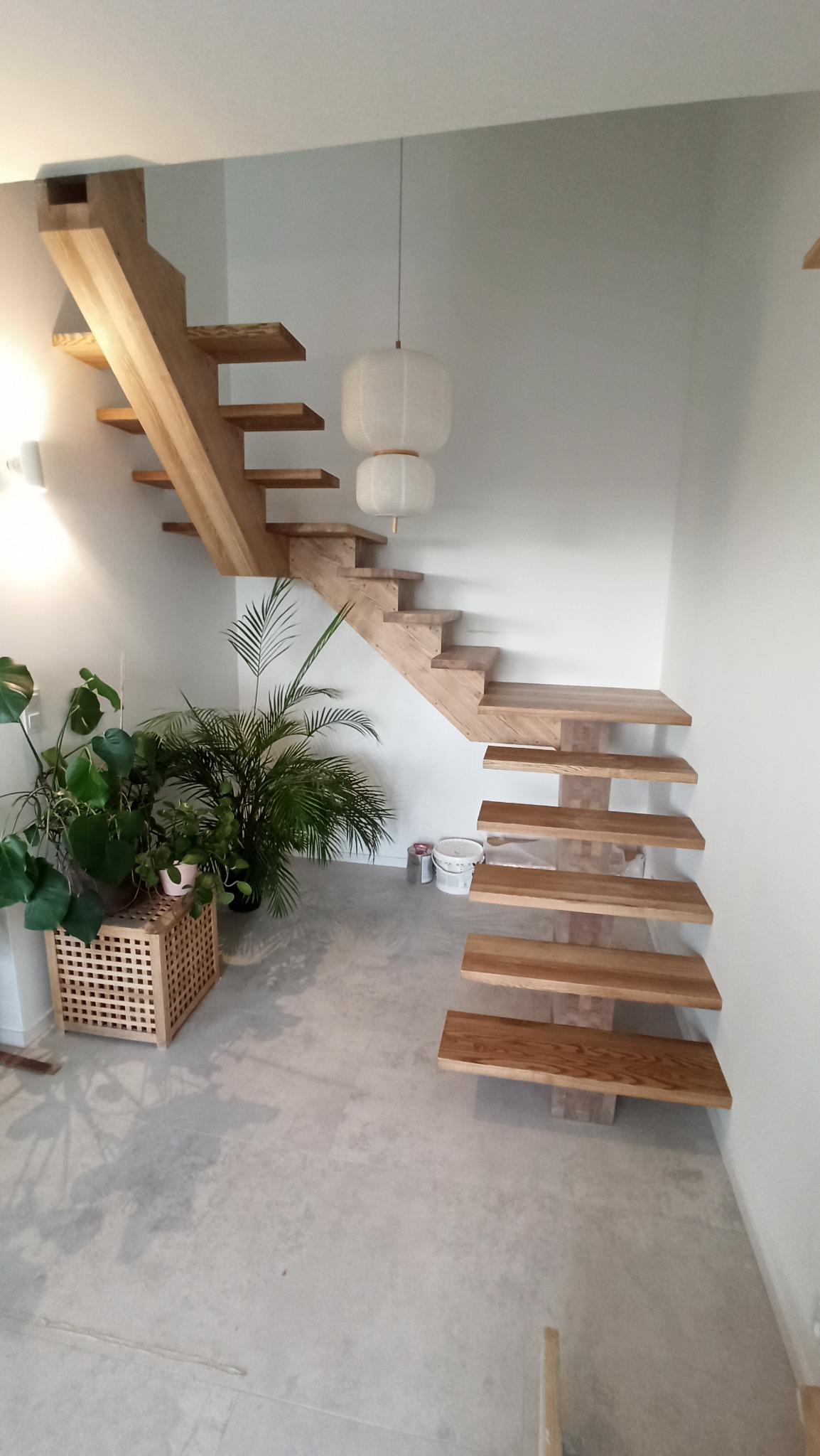 Minimalistyczne, drewniane schody wspornikowe z jasnego drewna, z roślinami doniczkowymi u podstawy, oświetlone wiszącą lampą w kształcie kuli.