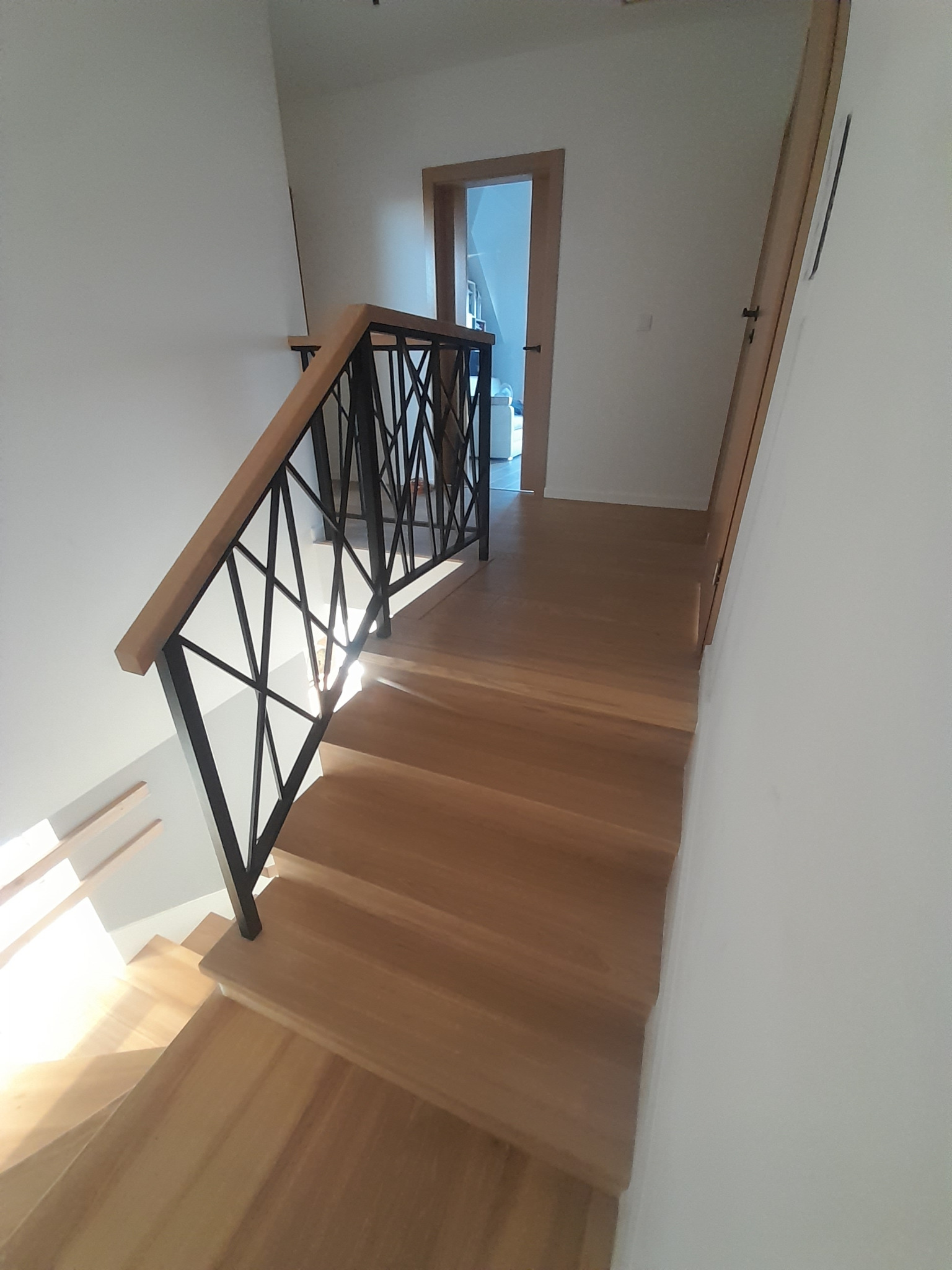 Jasne, drewniane stopnie schodów z czarną, geometryczną balustradą i drewnianą poręczą, prowadzące w górę do otwartych drzwi.