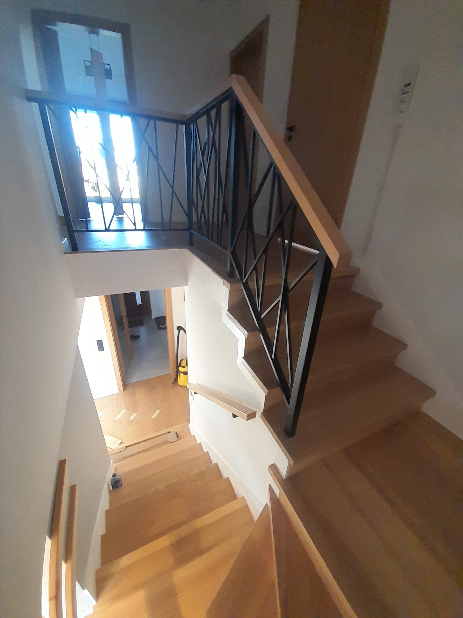 Widok z góry na drewniane schody z czarną, geometryczną balustradą i drewnianą poręczą, prowadzące w dół; w tle widoczne wnętrze domu z otwartymi drzwiami i odkurzaczem.