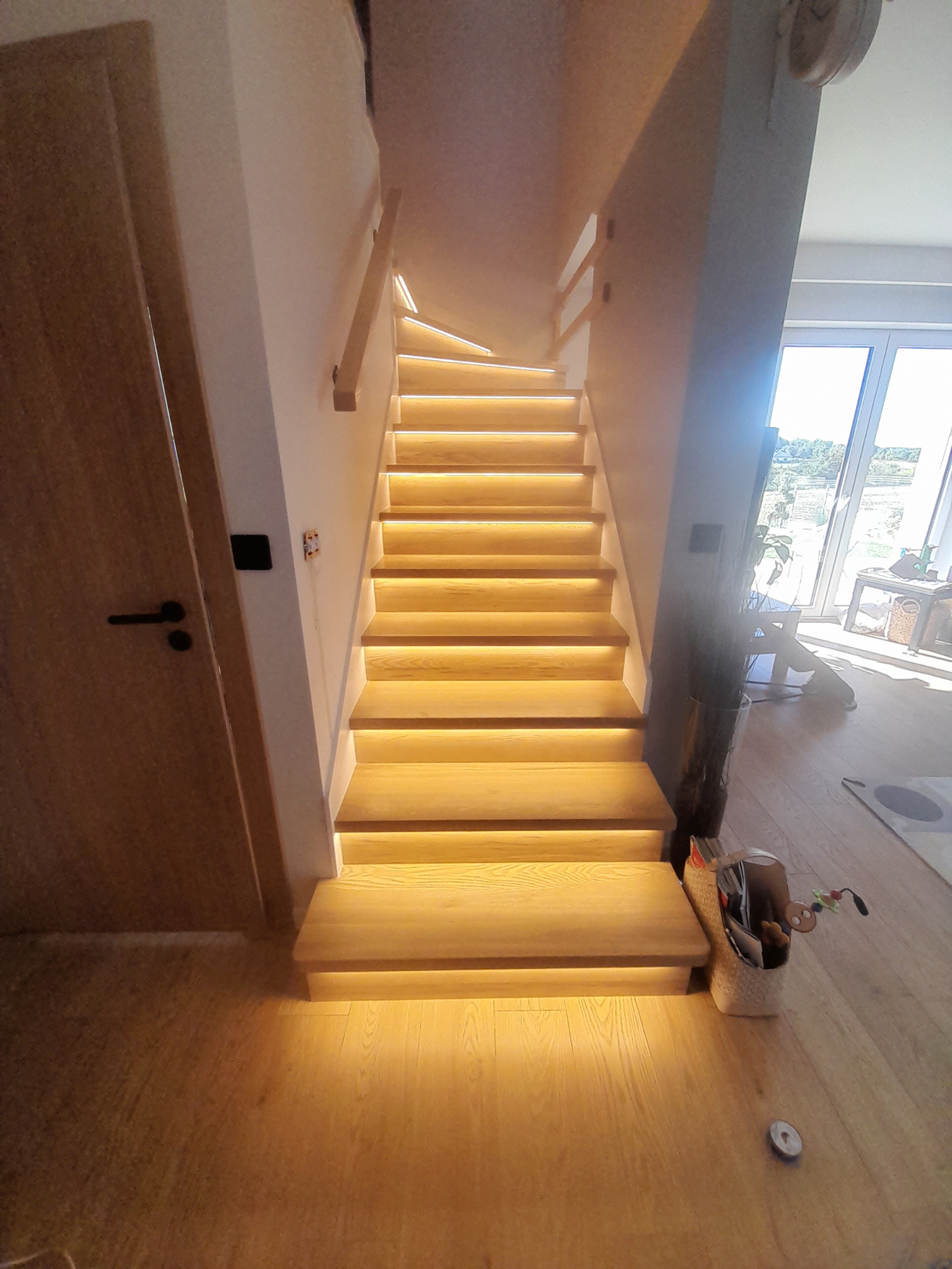 Nowoczesne schody z podświetleniem LED, drewniane stopnie, jasne wnętrze, widok z dołu, drzwi po lewej stronie, koszyk z zabawkami u podstawy schodów.