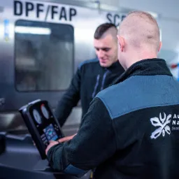 Dwóch mechaników obsługuje maszynę do czyszczenia filtrów DPF/FAP w warsztacie, widoczny panel sterowania i logo firmy na ubraniu jednego z nich.