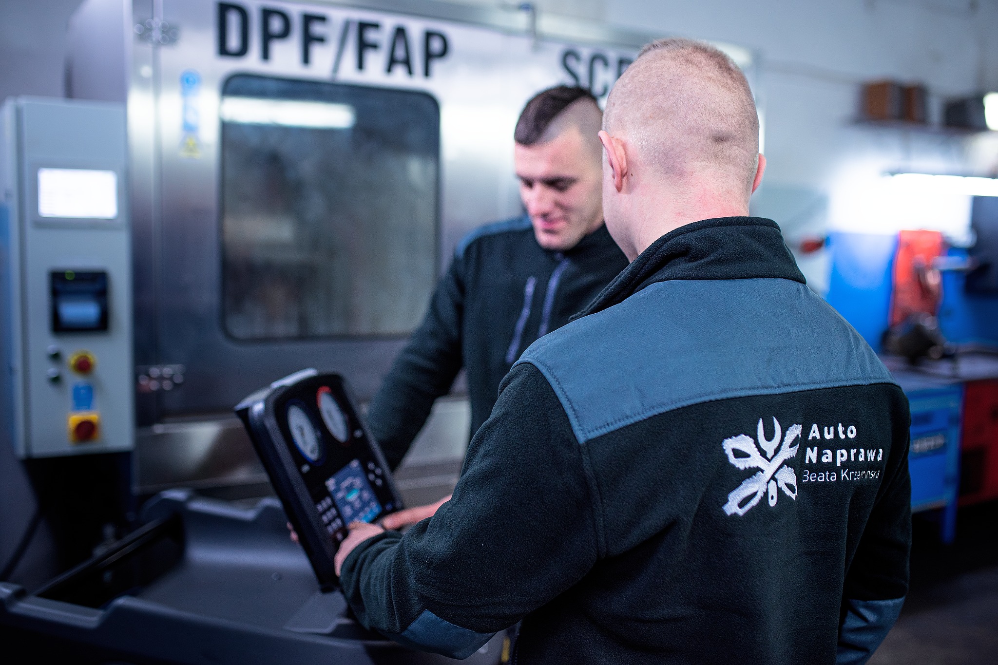 Dwóch mechaników obsługuje maszynę do czyszczenia filtrów DPF/FAP w warsztacie, widoczny panel sterowania i logo firmy na ubraniu jednego z nich.