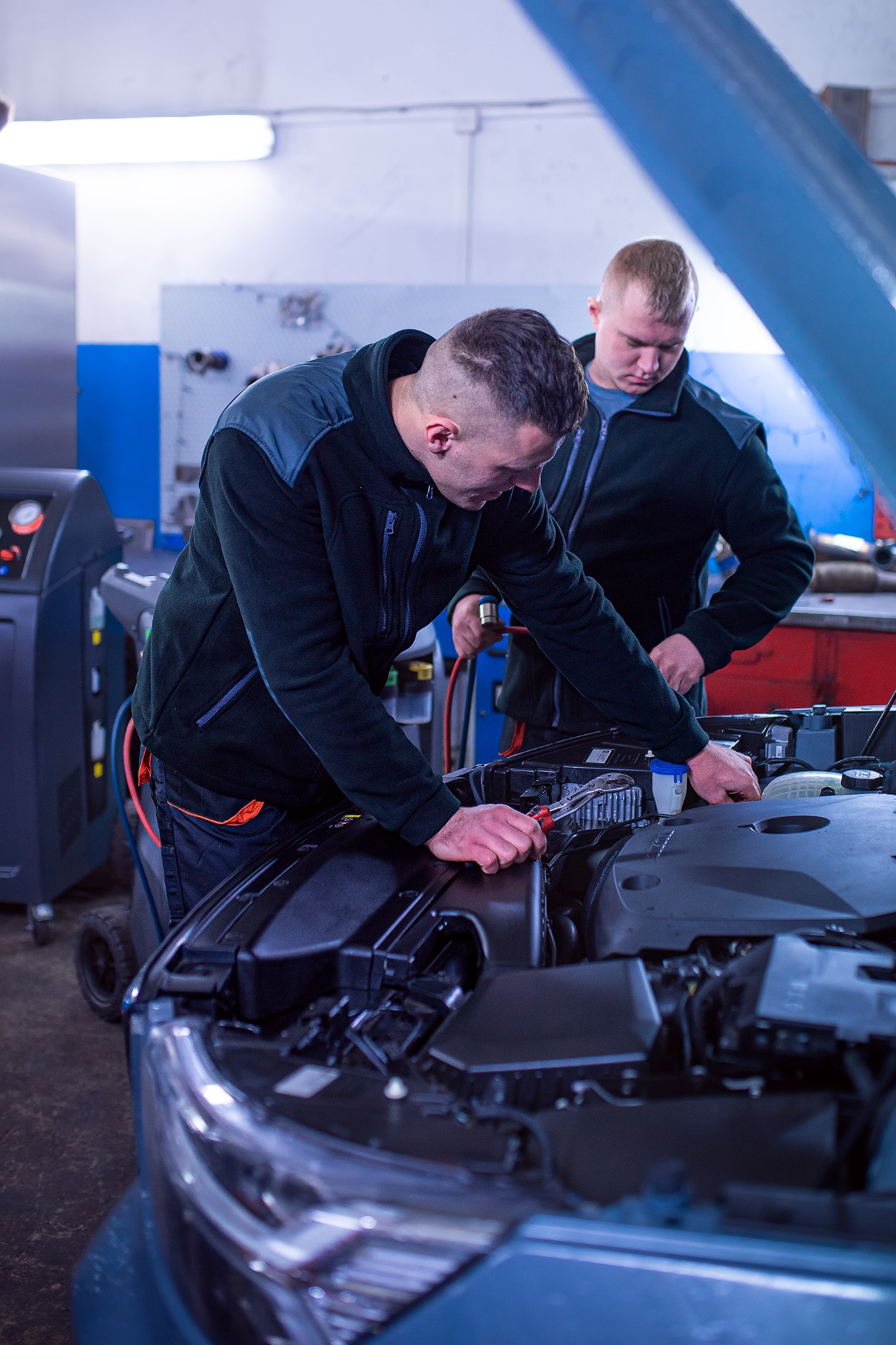Dwóch mechaników pochylonych nad otwartą maską samochodu w warsztacie, jeden z kluczem nastawnym w dłoni, drugi sprawdza akumulator, w tle maszyna do klimatyzacji.