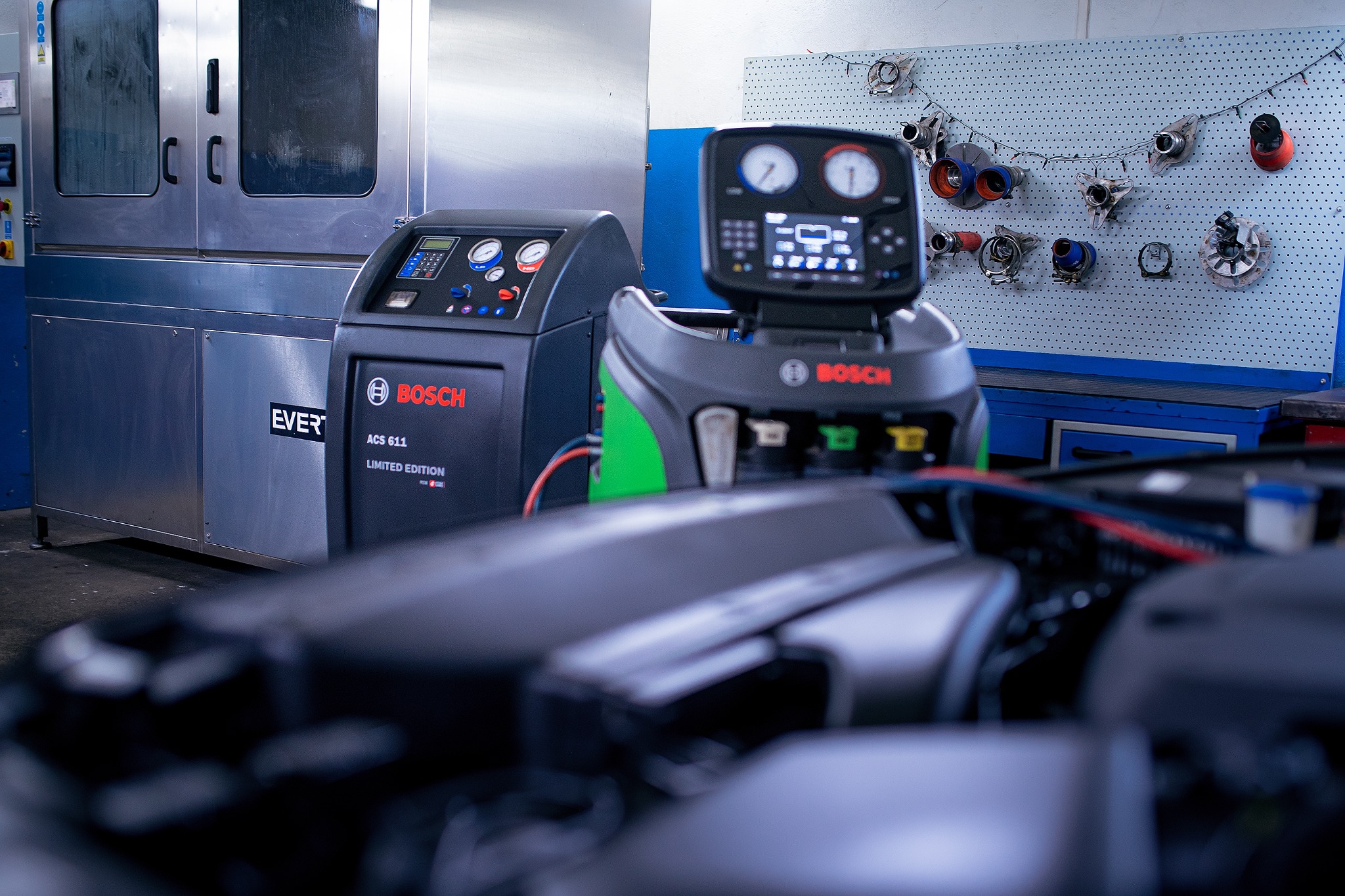 Dwa urządzenia Bosch ACS 611 Limited Edition do obsługi klimatyzacji samochodowej w warsztacie, z widoczną otwartą maską samochodu na pierwszym planie i ścianą z narzędziami w tle.