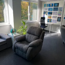 Wnętrze gabinetu z szarym, rozkładanym fotelem, rośliną doniczkową, turkusowym wentylatorem i ścianą z grafikami przedstawiającymi mózg.